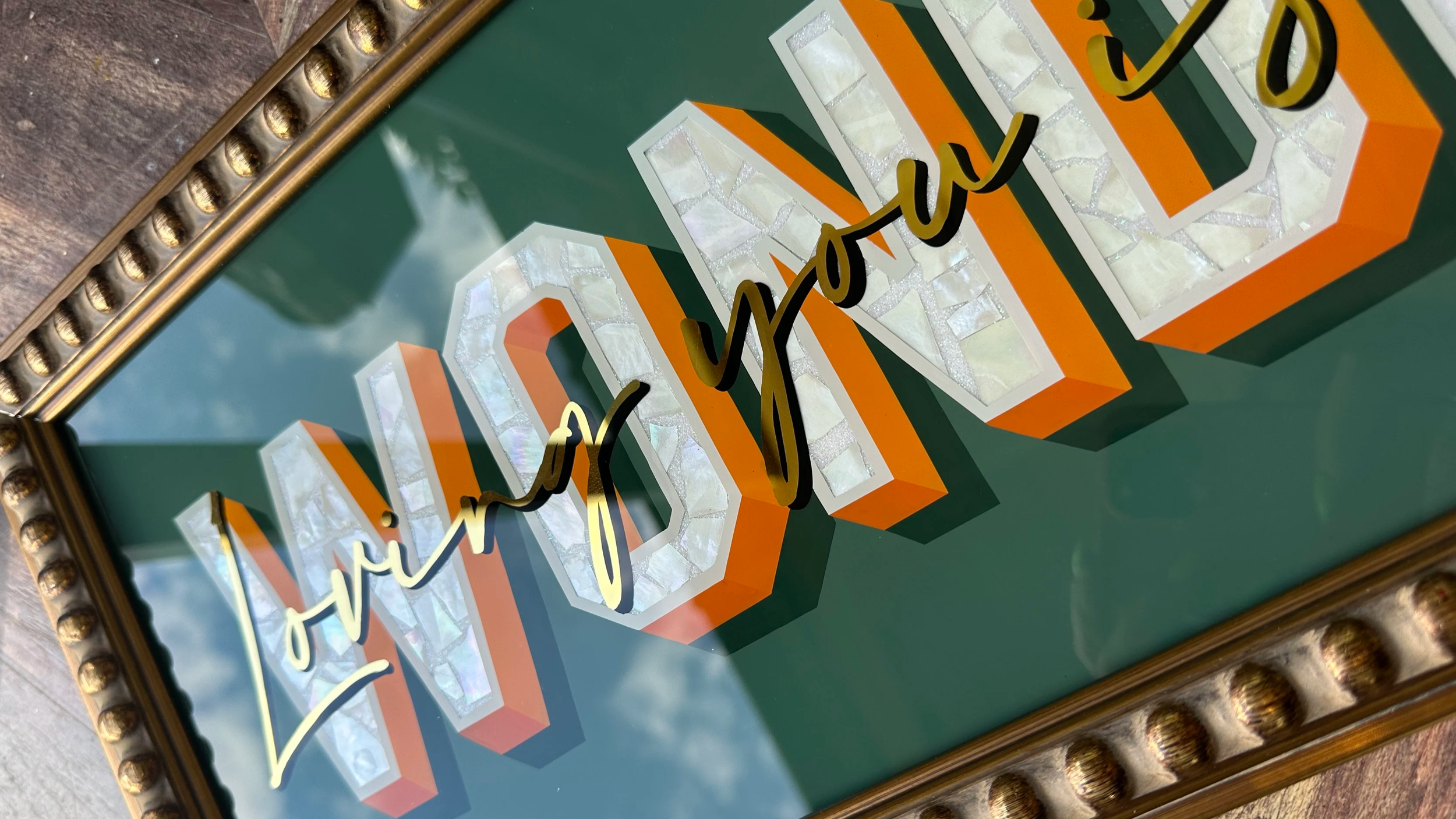 Decorative sign with large 3D block letters spelling 'LOVE' in white and orange, with cursive black and yellow script overlay reading 'love points', in a gold-finished ornate frame, reflecting a cloudy sky.