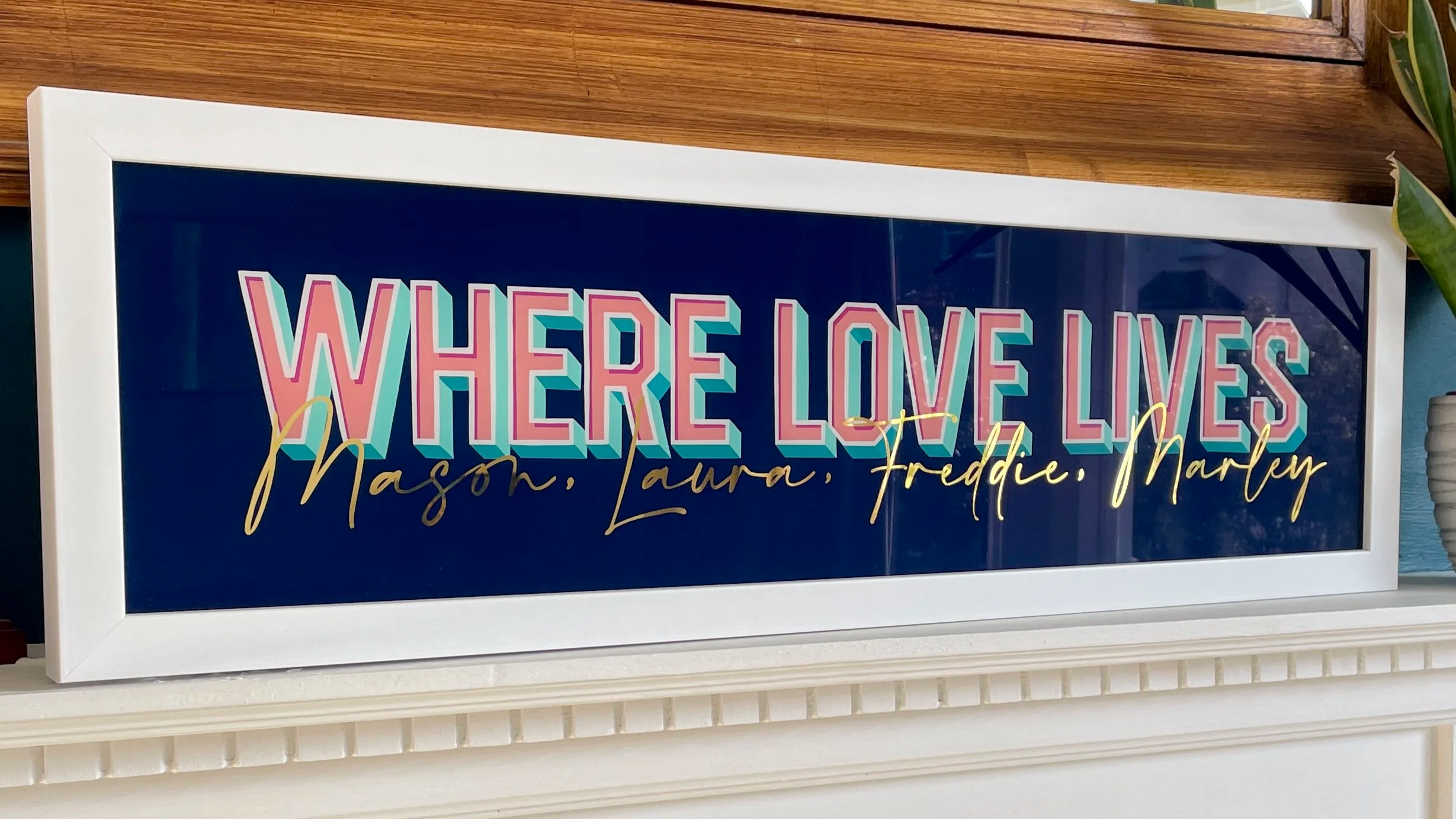 Decorative sign with the phrase 'Where Love Lives' in colorful, bold letters, and names Mason, Laura, Freddie, and Marley written in gold cursive below.