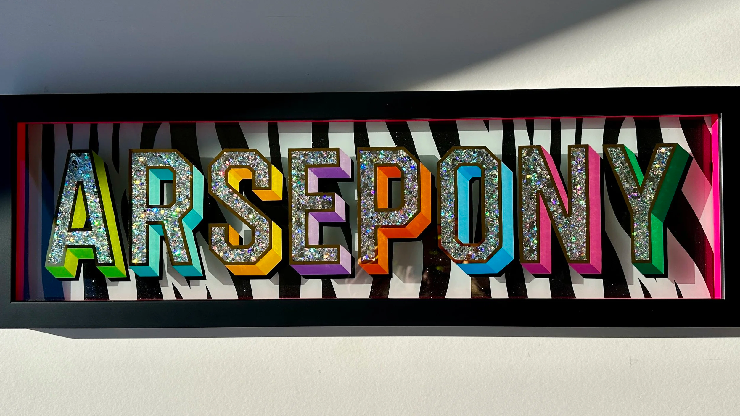 Colorful sign spelling 'ARSEYON' with glittery letters and rainbow outlines, mounted on a zebra print background.