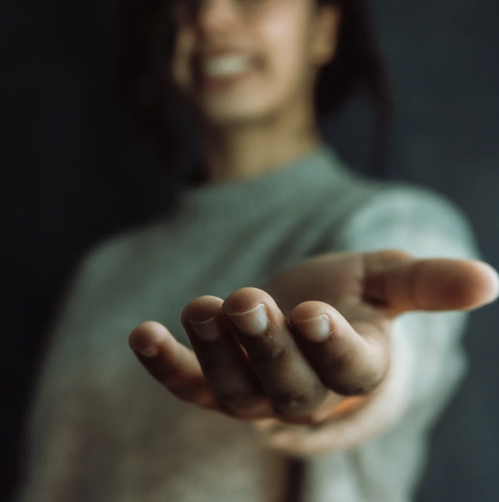 Close-up of a smiling woman extending her hand toward the camera, with a dark background.