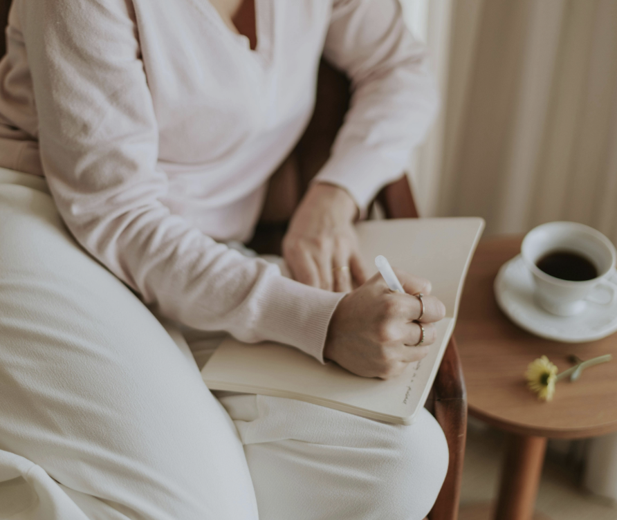 Person sitting on a chair writing in a notebook with a pen, a cup of coffee on a small table, and a yellow flower nearby.