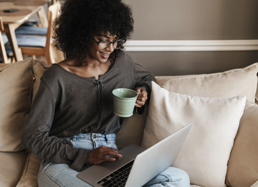 Woman with curly hair and glasses using a laptop while sitting on a beige couch, holding a green mug.