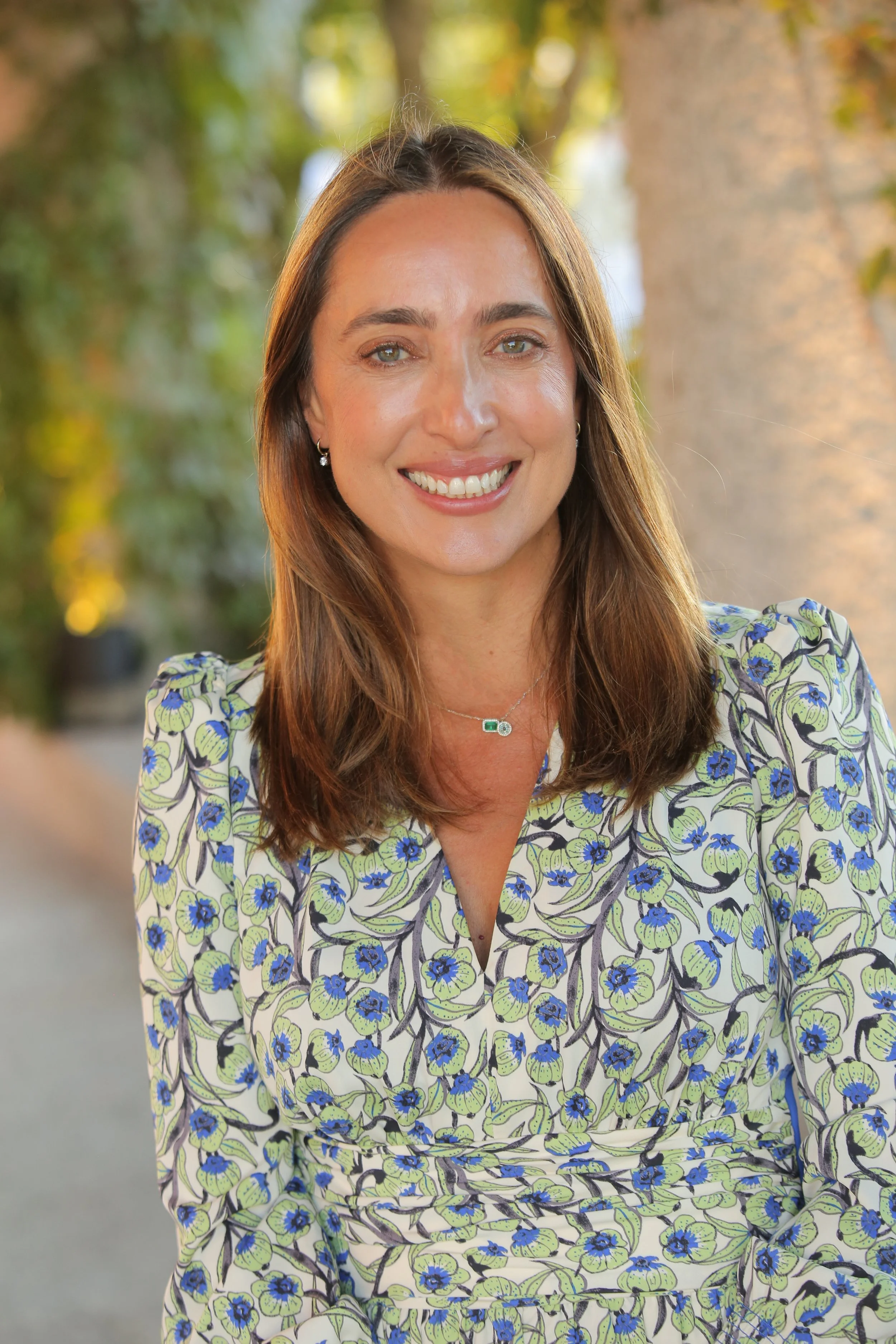 A woman with long brown hair and blue eyes, smiling outdoors, wearing a colorful floral dress with green, blue, and black patterns, and a silver necklace with a green gemstone.