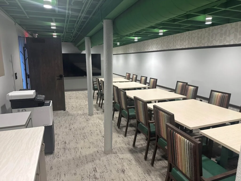 A conference room with multiple tables and chairs, a large television on the wall, and a green ceiling with visible ductwork.