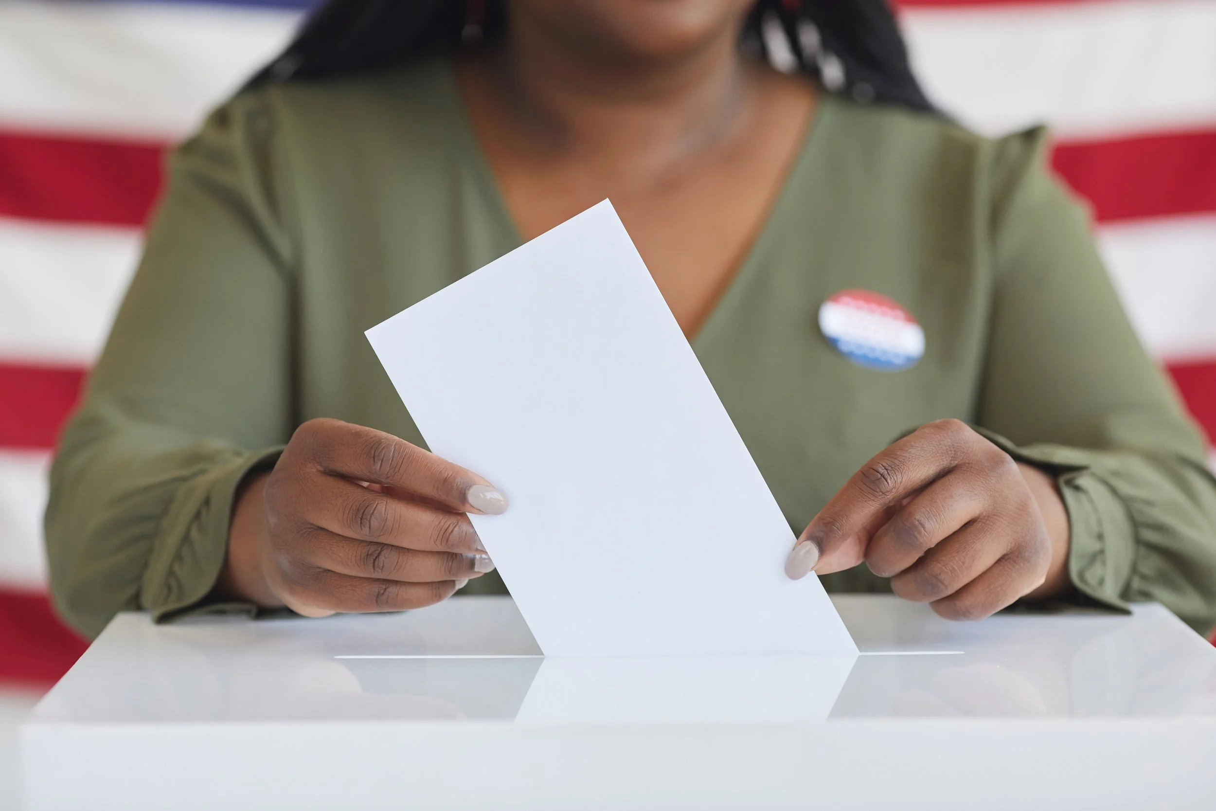A person casting a vote by inserting a blank piece of paper into a ballot box, with an American flag in the background.
