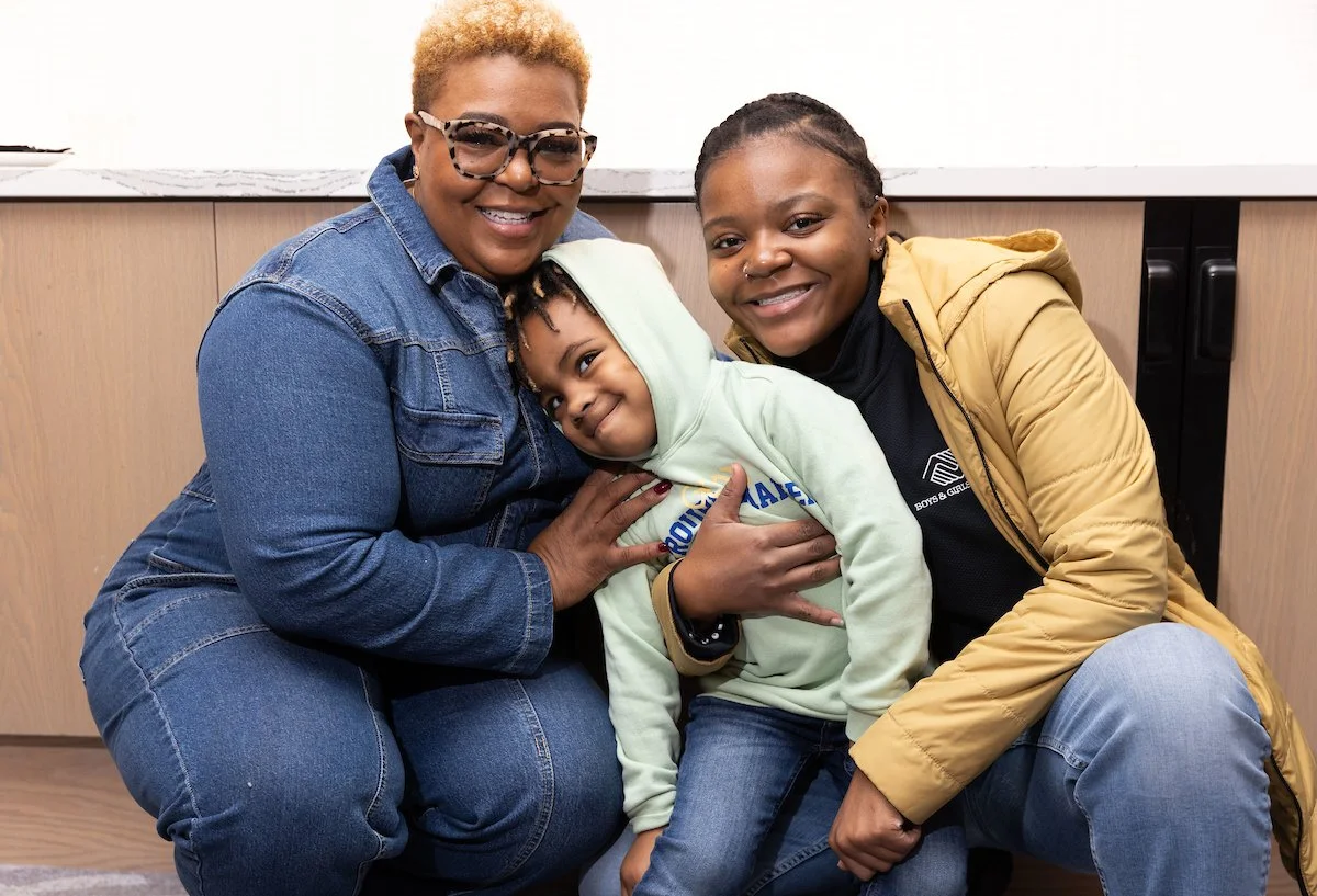 Three smiling females, a woman with short blonde hair, glasses, and a denim jacket, a woman with braided hair and a yellow jacket, and a young girl in a light green hoodie, in an indoor setting with a wooden cabinet in the background.