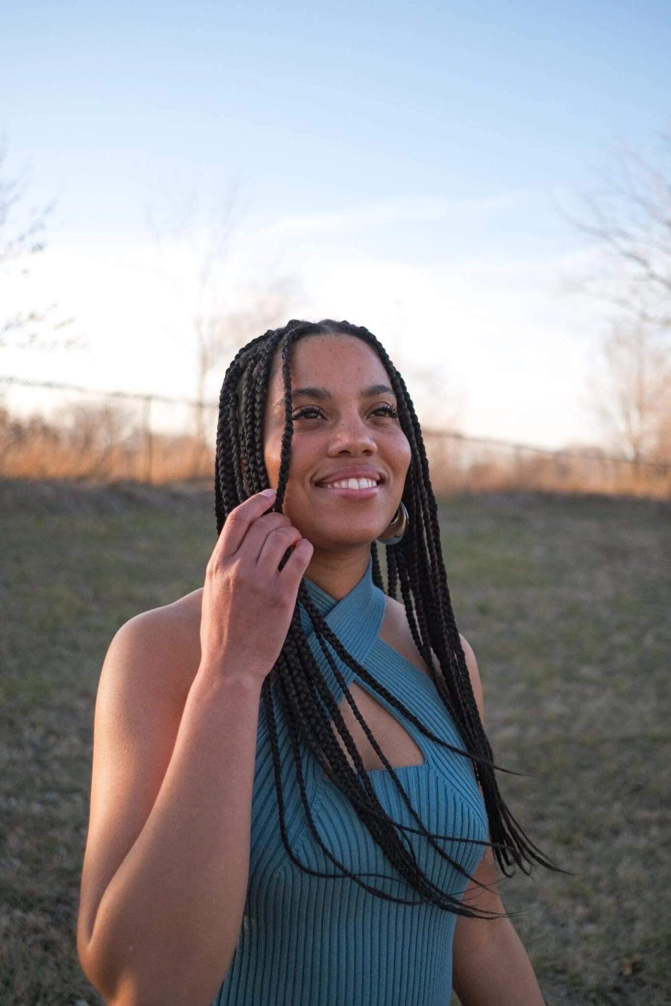 A smiling woman with braided hair in a blue sleeveless dress standing outdoors during sunset.