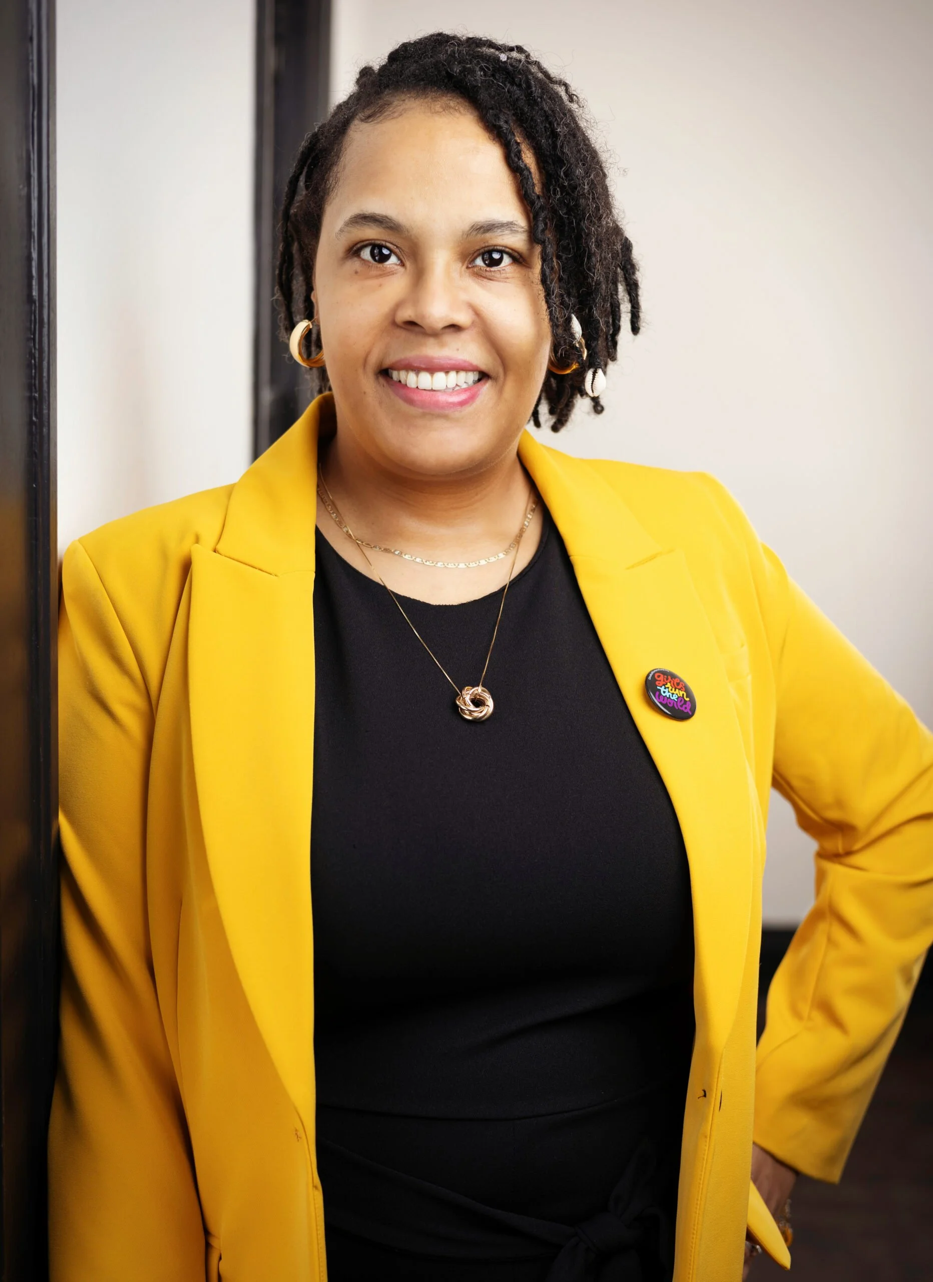 A woman with natural curly hair wearing a yellow blazer over a black top, accessorized with gold jewelry and a colorful button pin, standing confidently near a wall.