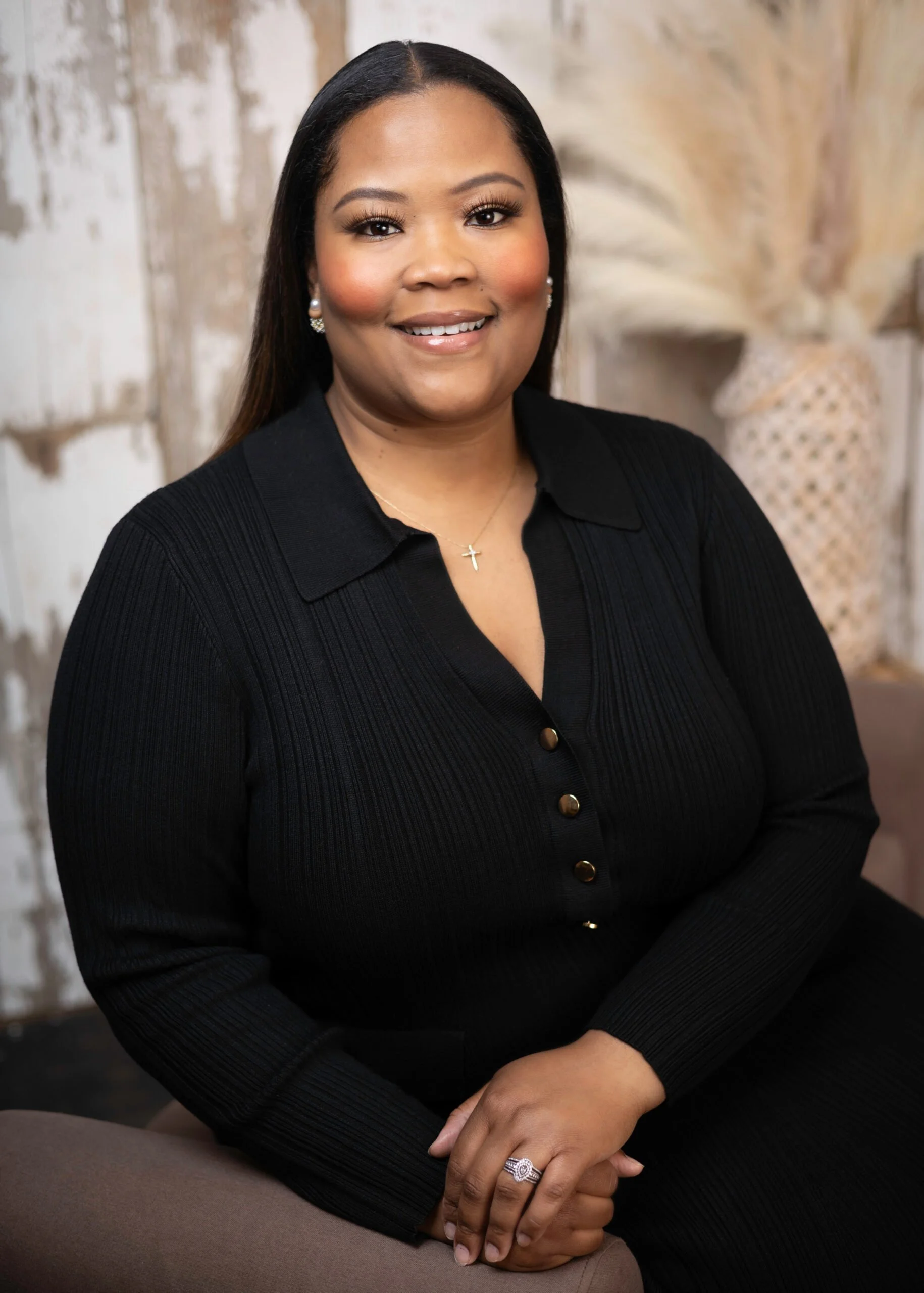 A woman with long dark hair, smiling, wearing a black button-up sweater, a cross necklace, earrings, and a ring, seated with hands resting on her lap, against a neutral textured background.
