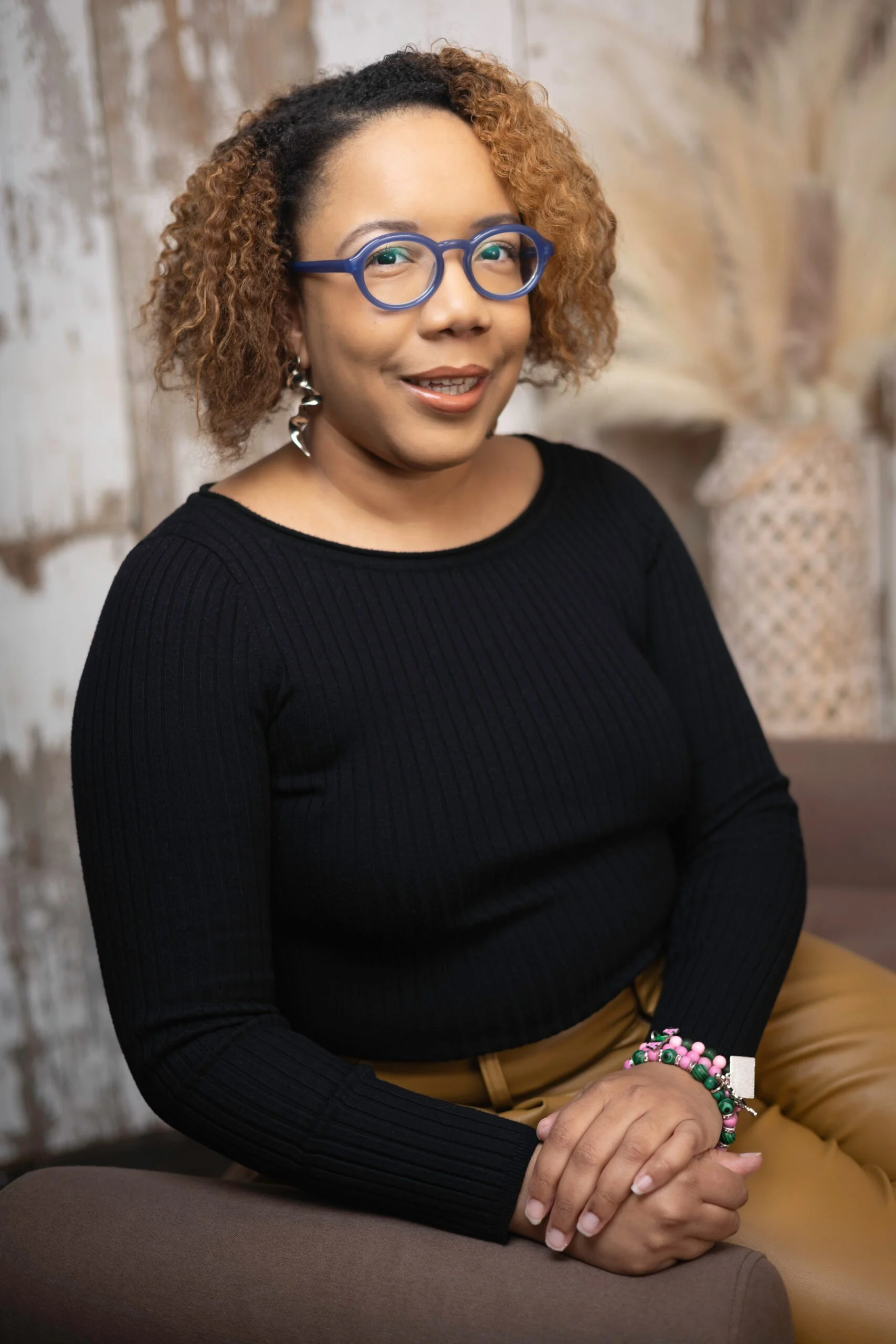 A woman with glasses and earrings wearing a black ribbed sweater and tan pants, sitting on a chair in front of a rustic wooden wall.
