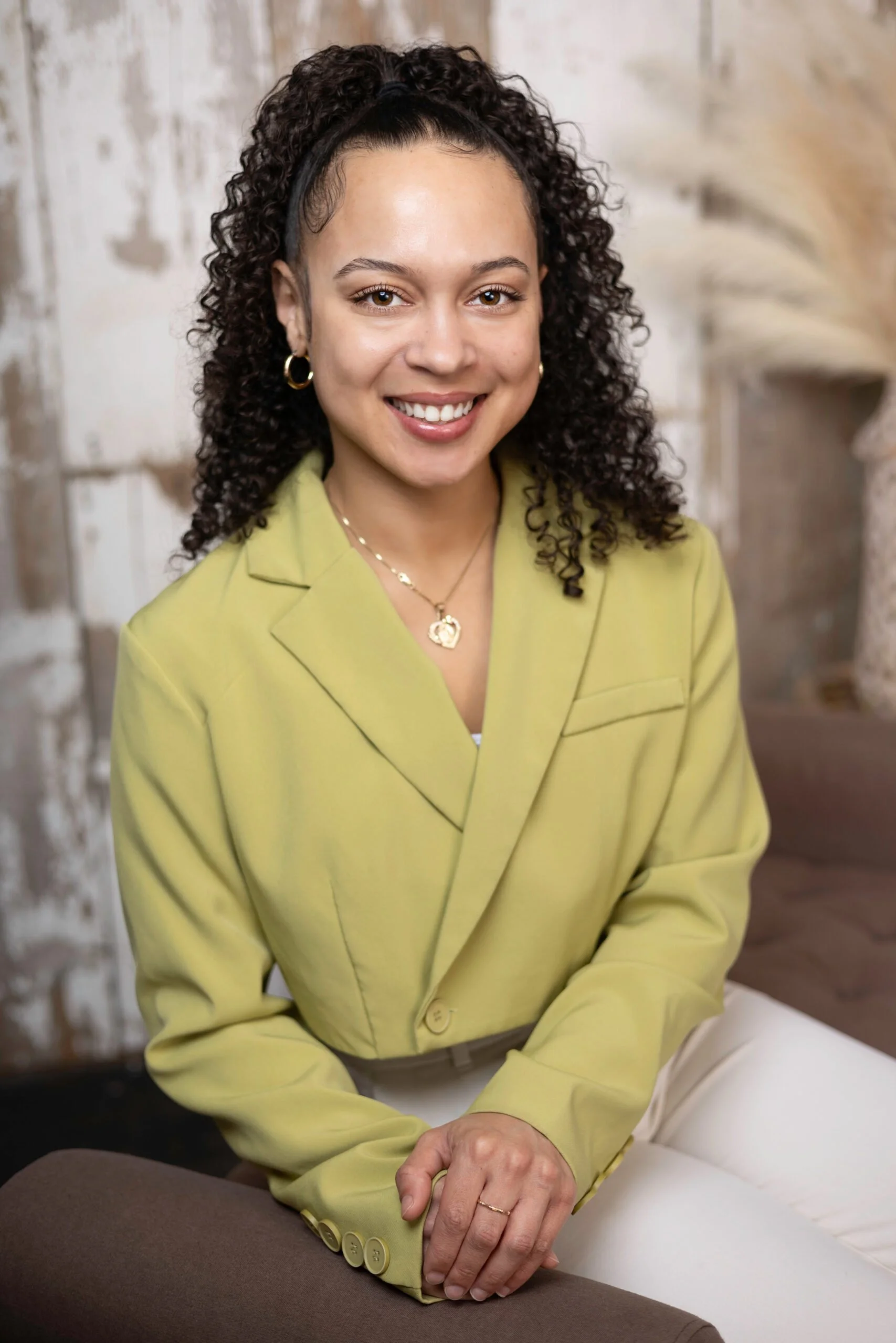 Smiling woman with curly hair wearing a lime green blazer, sitting on a brown armrest, with a blurred stone wall background.