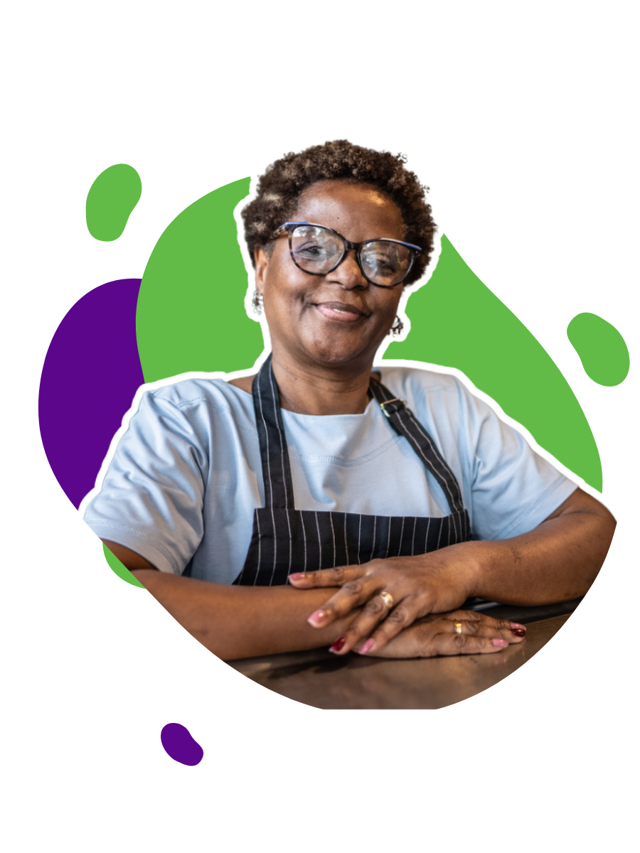 A smiling African American woman wearing glasses, a light blue shirt, and a dark striped apron, sitting with her forearms on a dark table, with arms crossed, in front of a colorful abstract background.