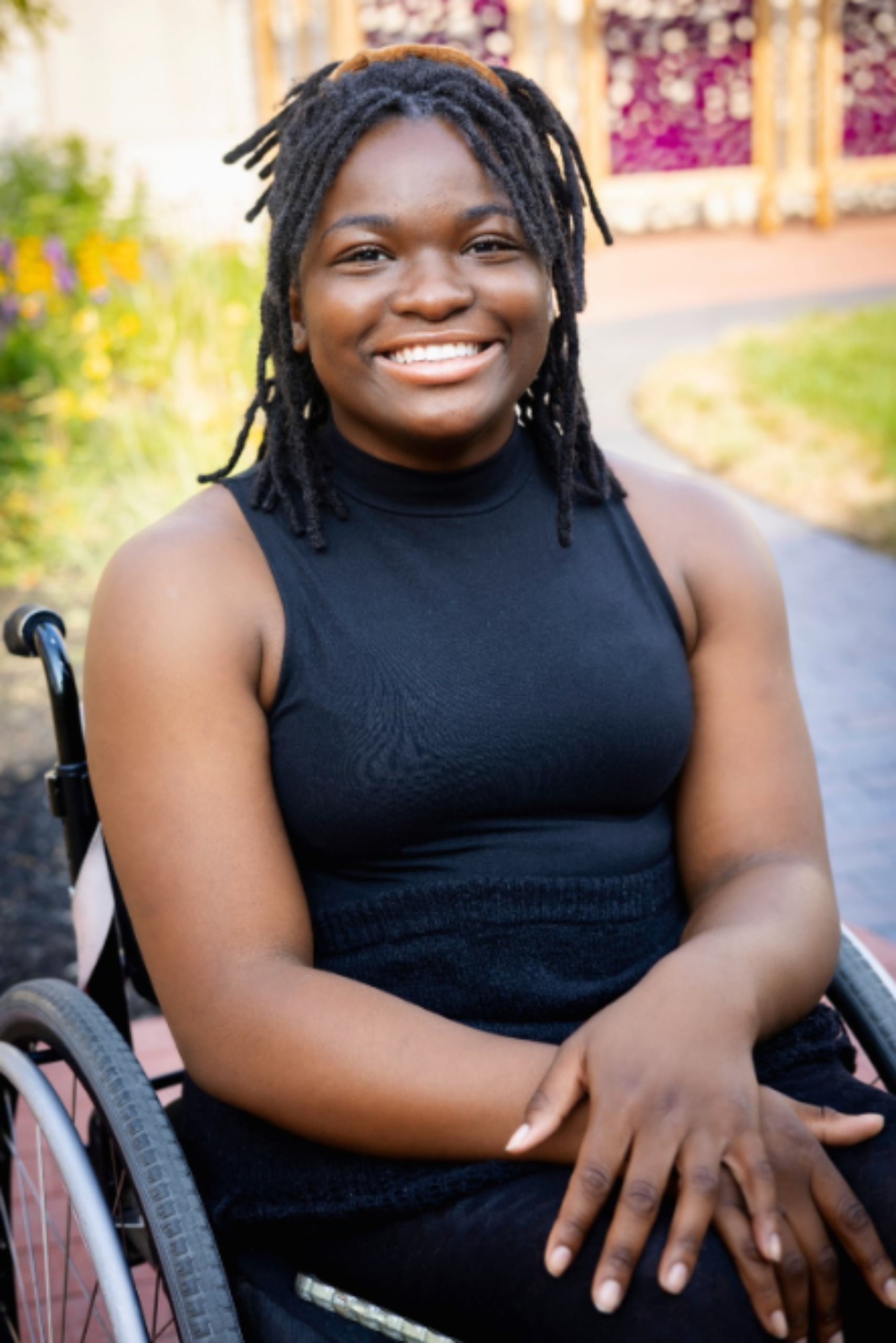 Smiling woman with dreadlocks sitting in a wheelchair outdoors, wearing a black sleeveless top, with colorful garden and pathway in the background.