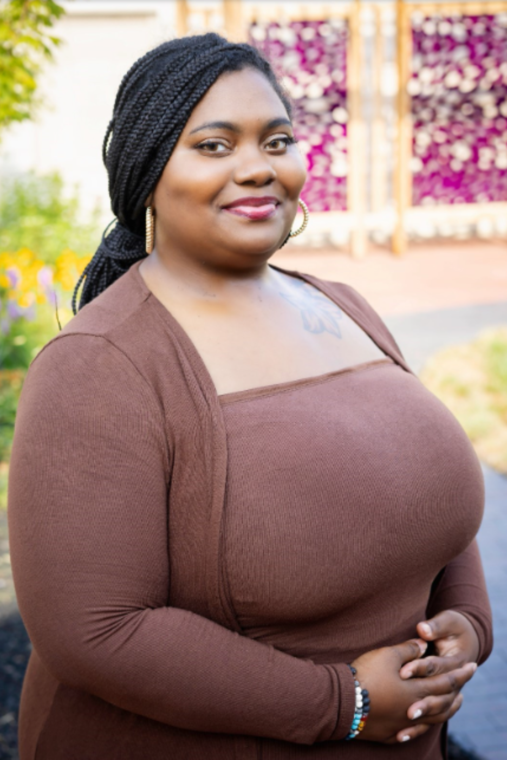 A woman with braided hair and gold hoop earrings smiling outdoors in a brown long-sleeve top, with colorful flowers and a garden in the background.