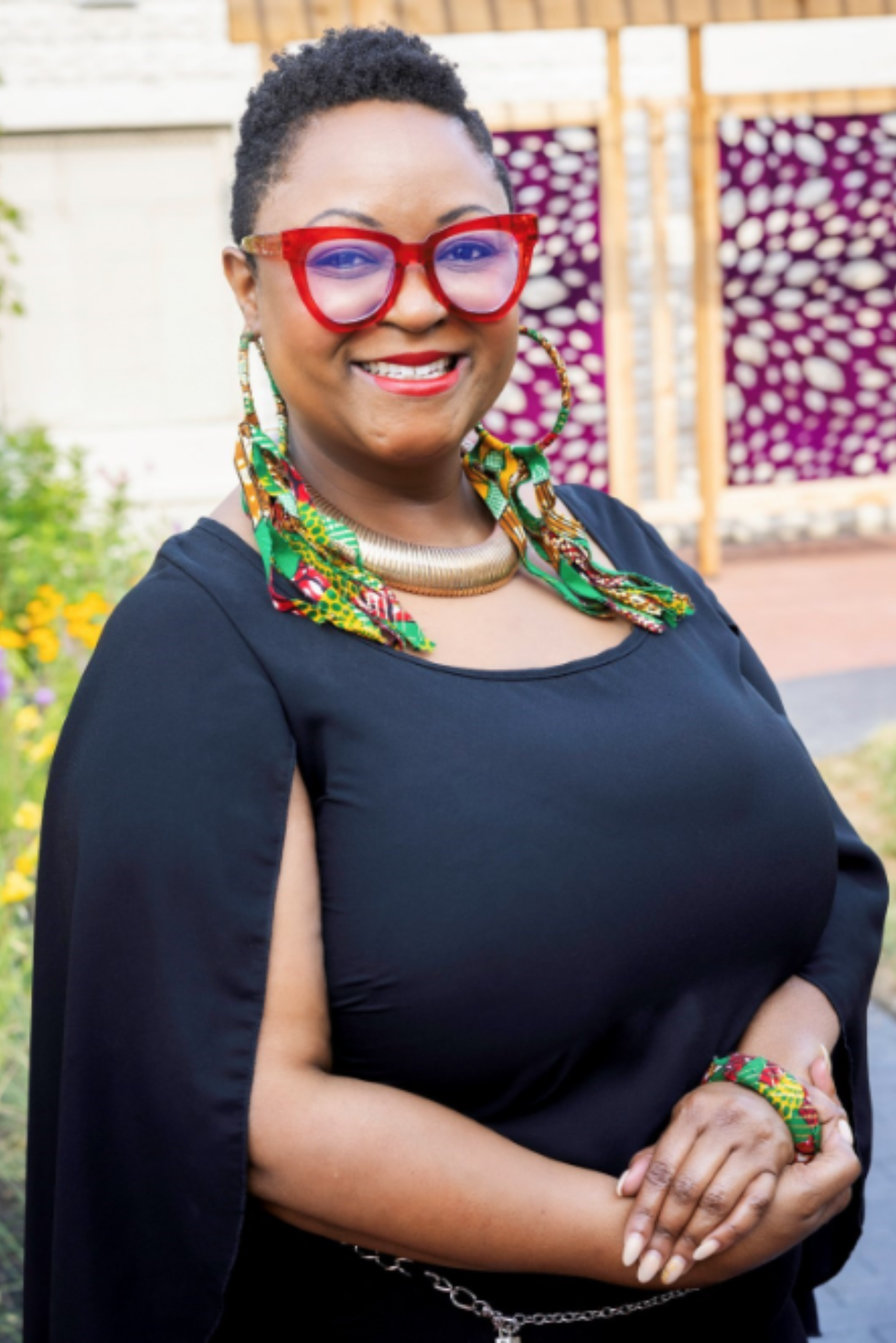 A woman with short curly hair wearing red glasses, colorful earrings, a gold choker, and a colorful scarf around her neck. She is dressed in a black top with cape-like sleeves and is standing outdoors with a flower garden and a decorative fence in the background, smiling at the camera.