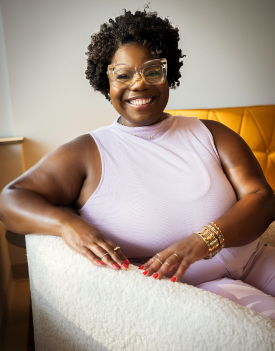 A smiling woman with glasses and curly hair sitting on a white armchair indoors, wearing a light purple sleeveless top and gold jewelry.