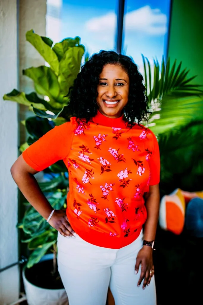 Smiling woman with curly black hair wearing an orange floral top and white pants, standing indoors with large green plants and a bright window in the background.
