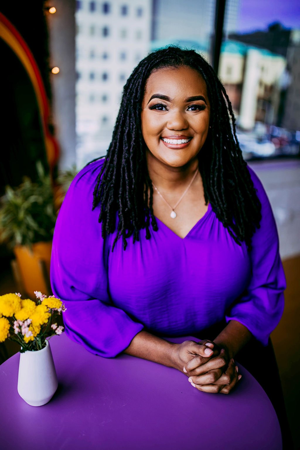 A smiling woman with long black dreadlocks wearing a purple top sitting at a purple table with a white vase of yellow flowers, indoors with a cityscape visible through the window in the background.