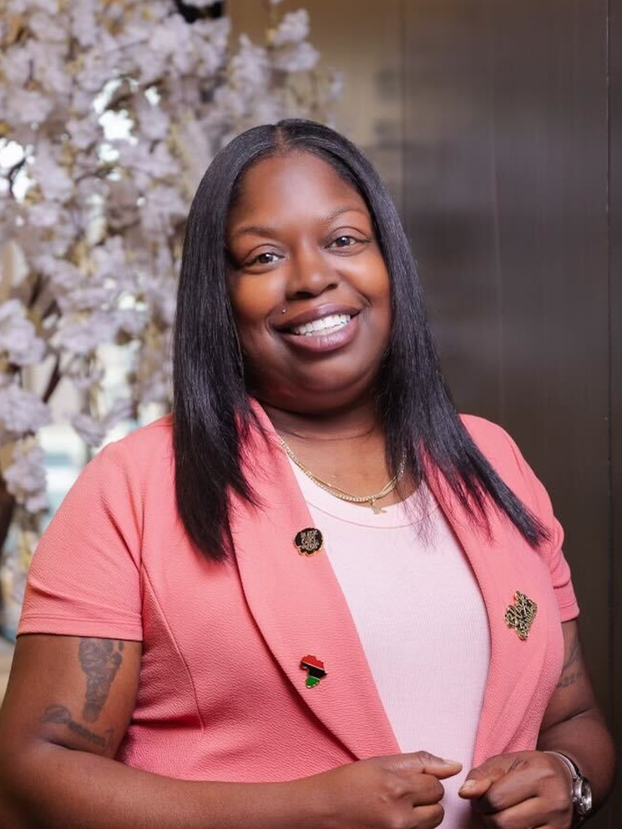 A woman with long black hair wearing a pink blazer with pins, a light pink shirt, and a gold necklace, standing in front of a floral background and smiling at the camera.