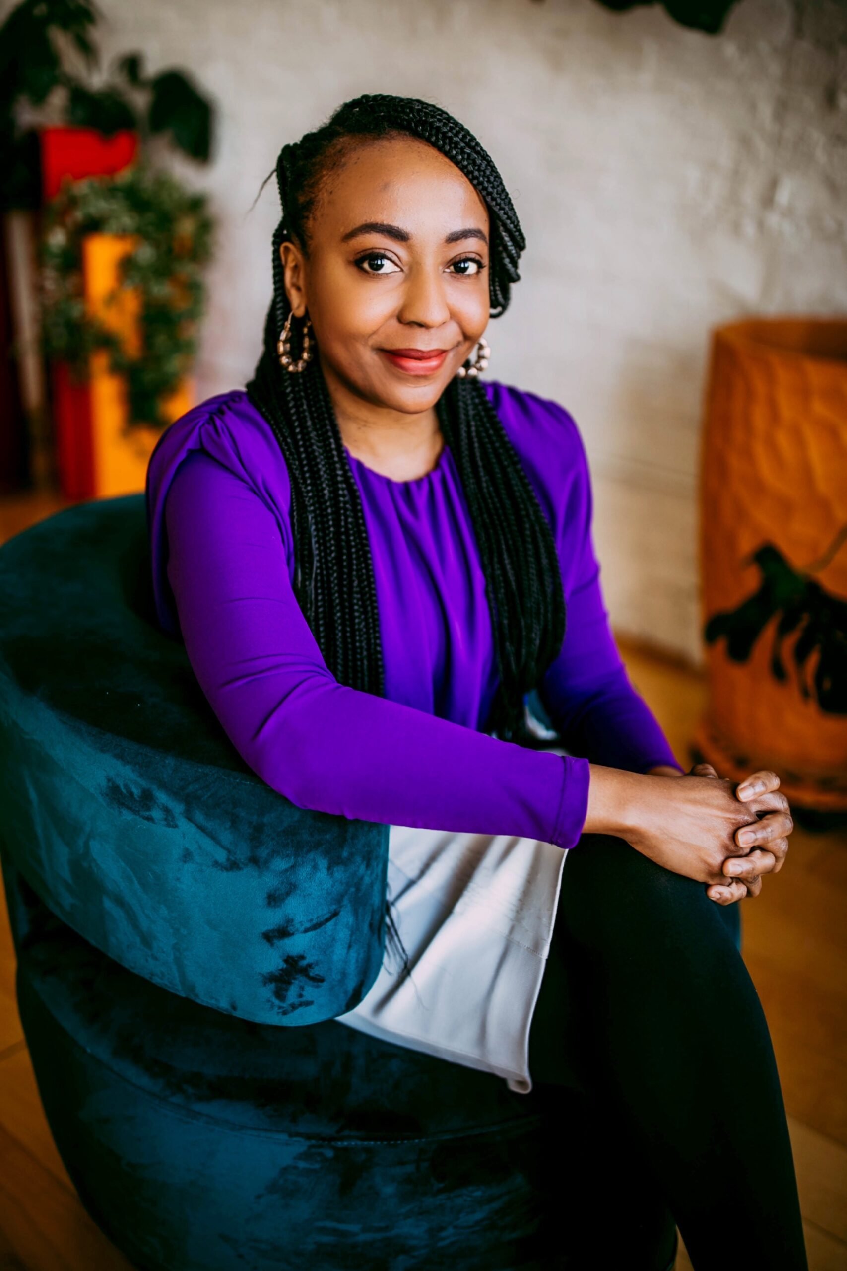 A woman with braided hair sitting on a dark green velvet chair wearing a purple blouse and earrings, indoors with a white brick wall and plant in the background.