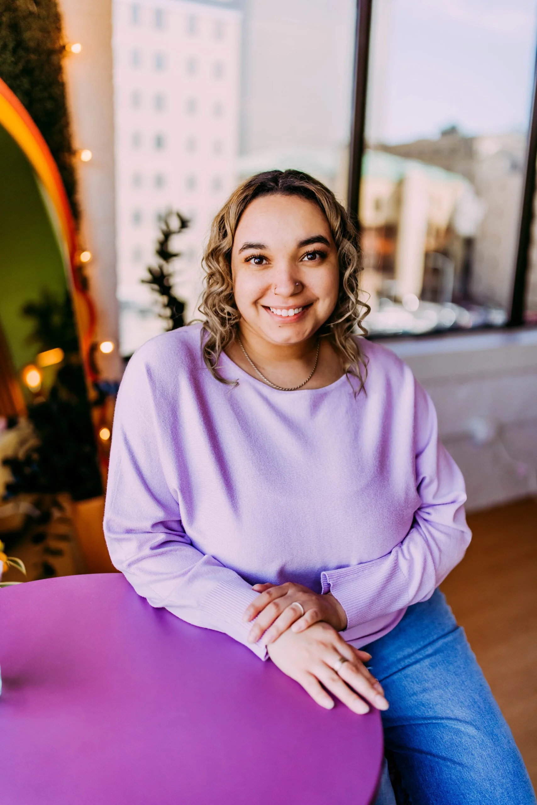 A young woman with curly hair and a nose piercing smiling while sitting at a purple table indoors, with a cityscape view through large windows behind her.