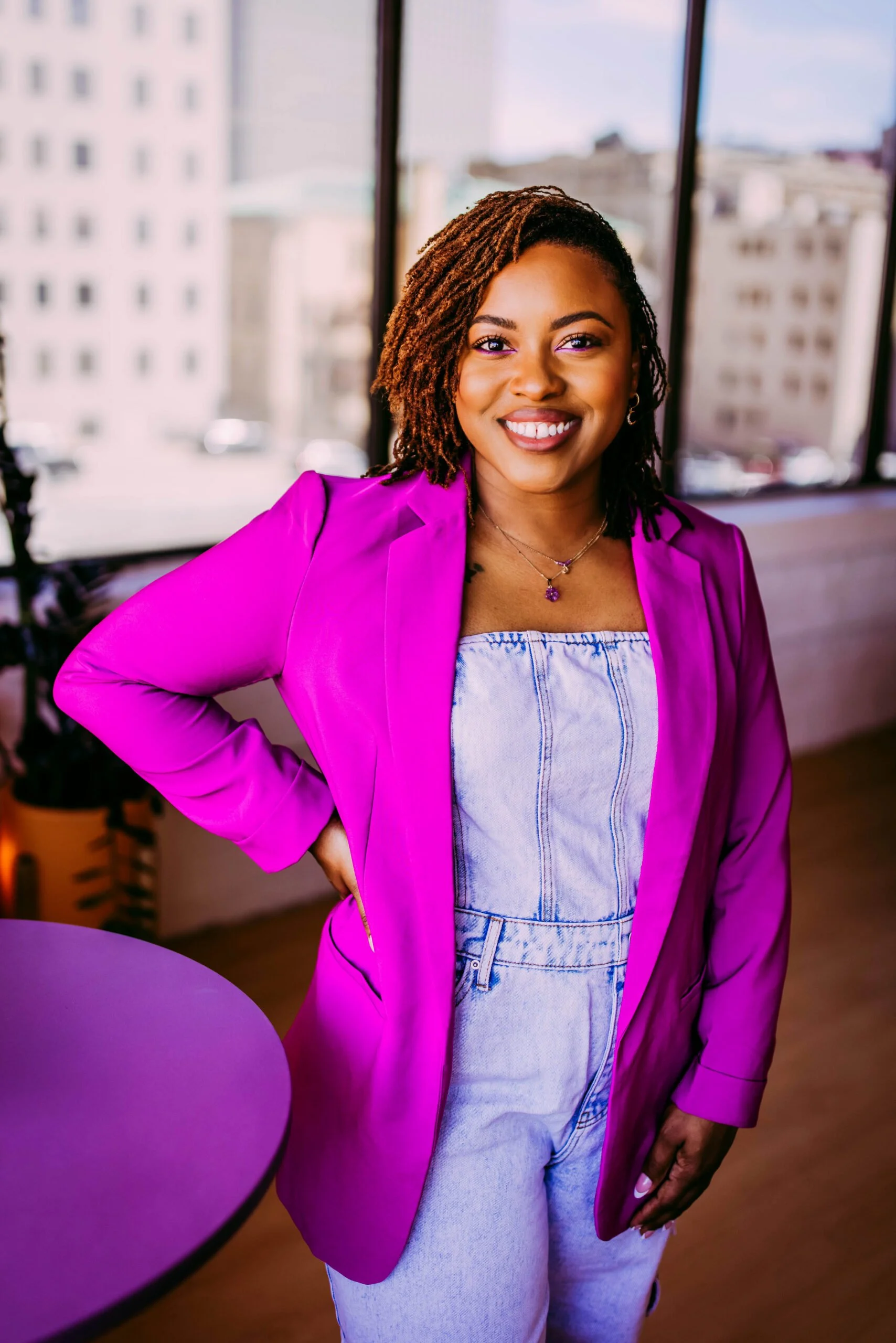 Smiling woman with short dreadlocks wearing a purple blazer and light denim jumpsuit, standing indoors next to a purple table, with cityscape view through large windows behind her.