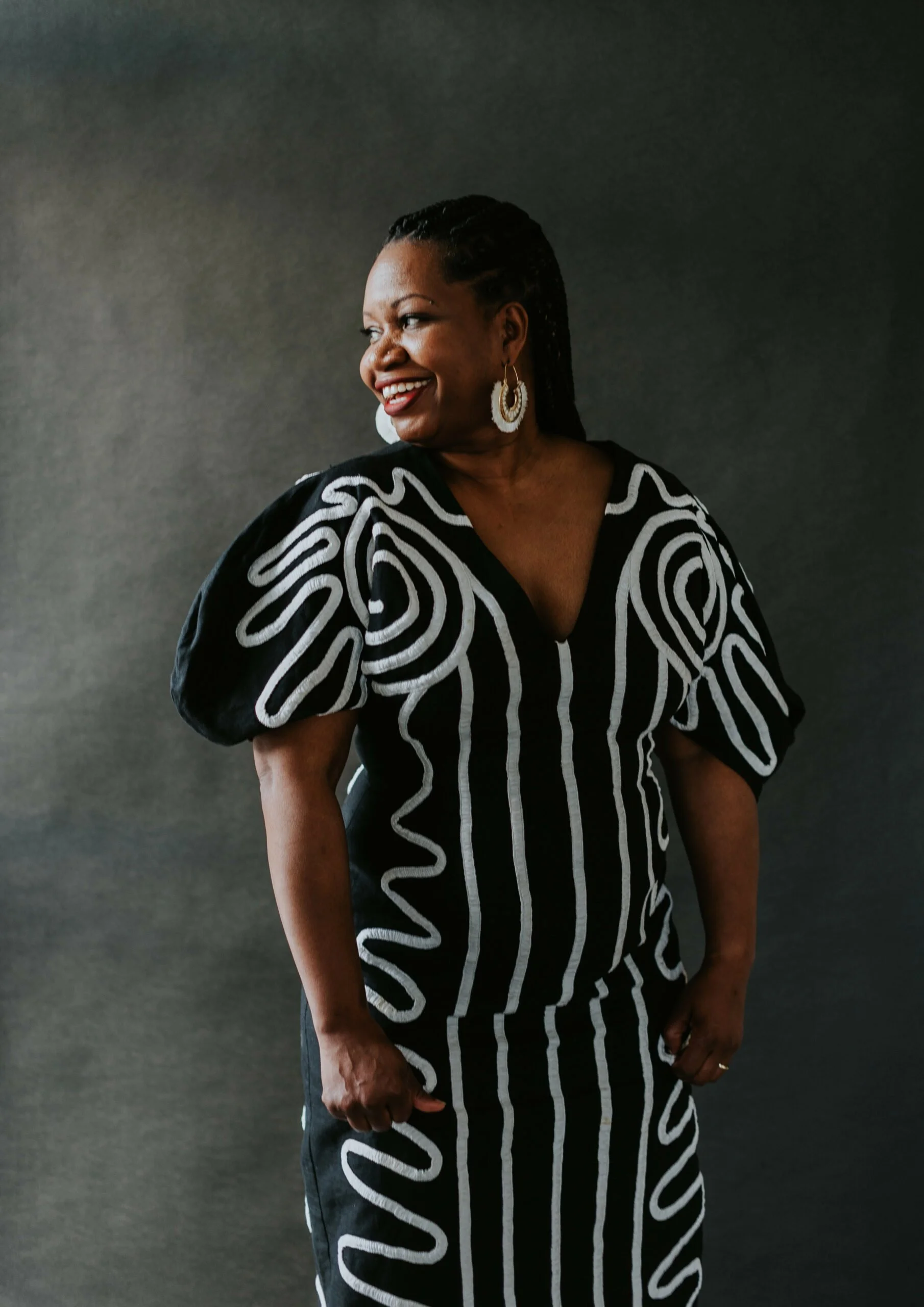 A woman wearing a black and white patterned dress with puffed sleeves, smiling and looking to her left against a dark gray background.