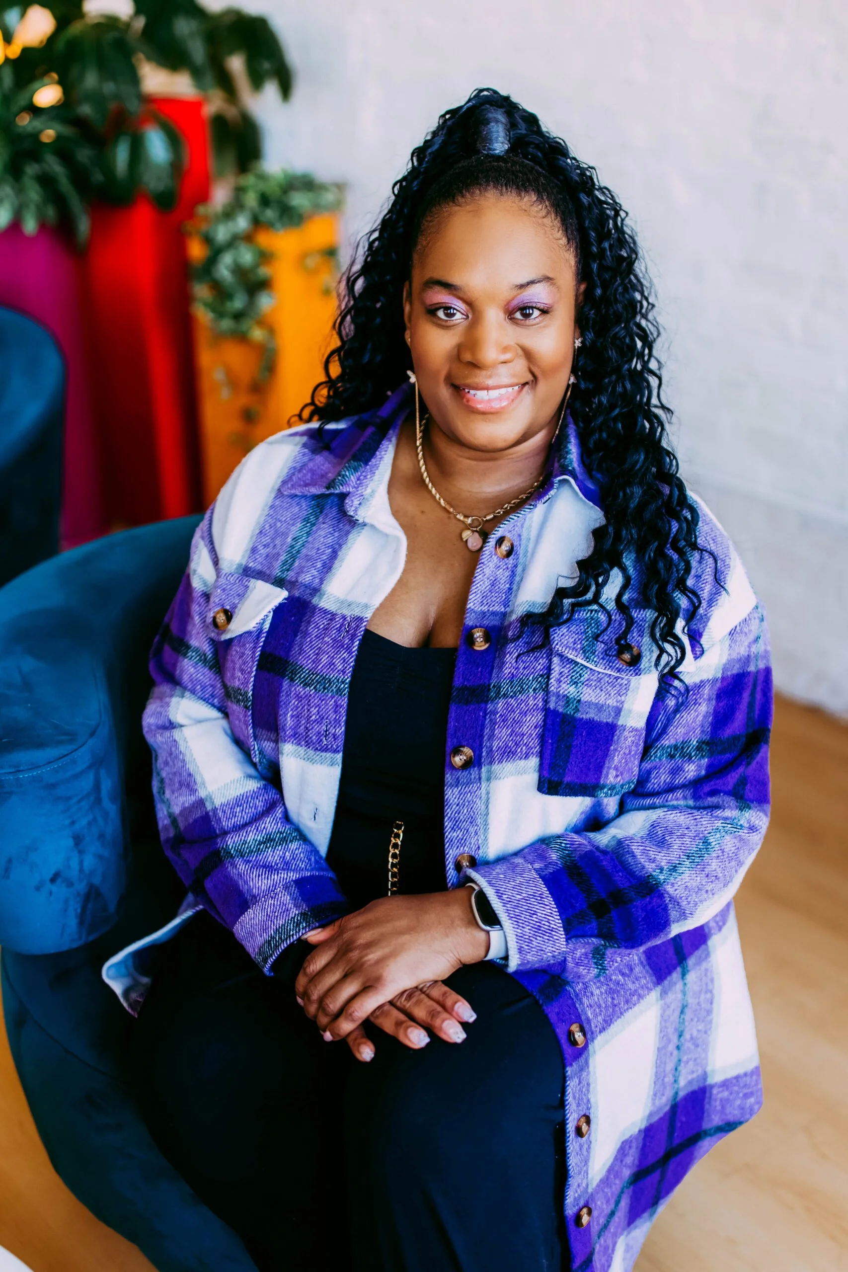 A woman with curly black hair in a high ponytail, wearing a purple and white plaid jacket with a black top, sitting on a blue chair in a brightly decorated room.