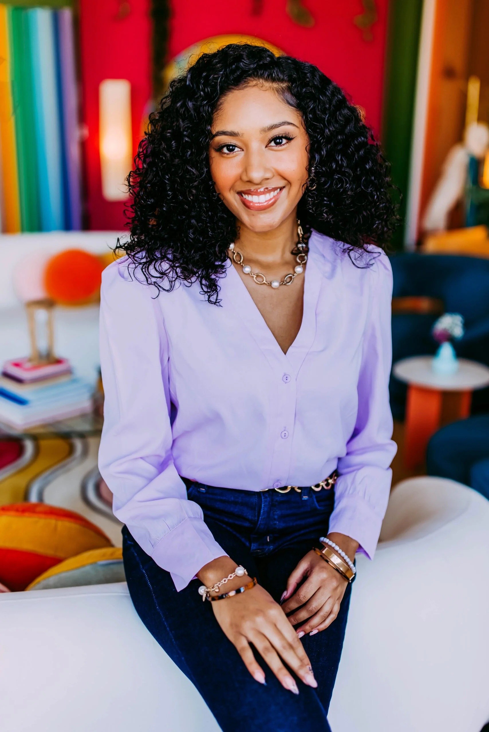 A woman with curly black hair smiling, wearing a light purple blouse, dark jeans, and jewelry, sitting in a colorful room with books, cushions, and décor.