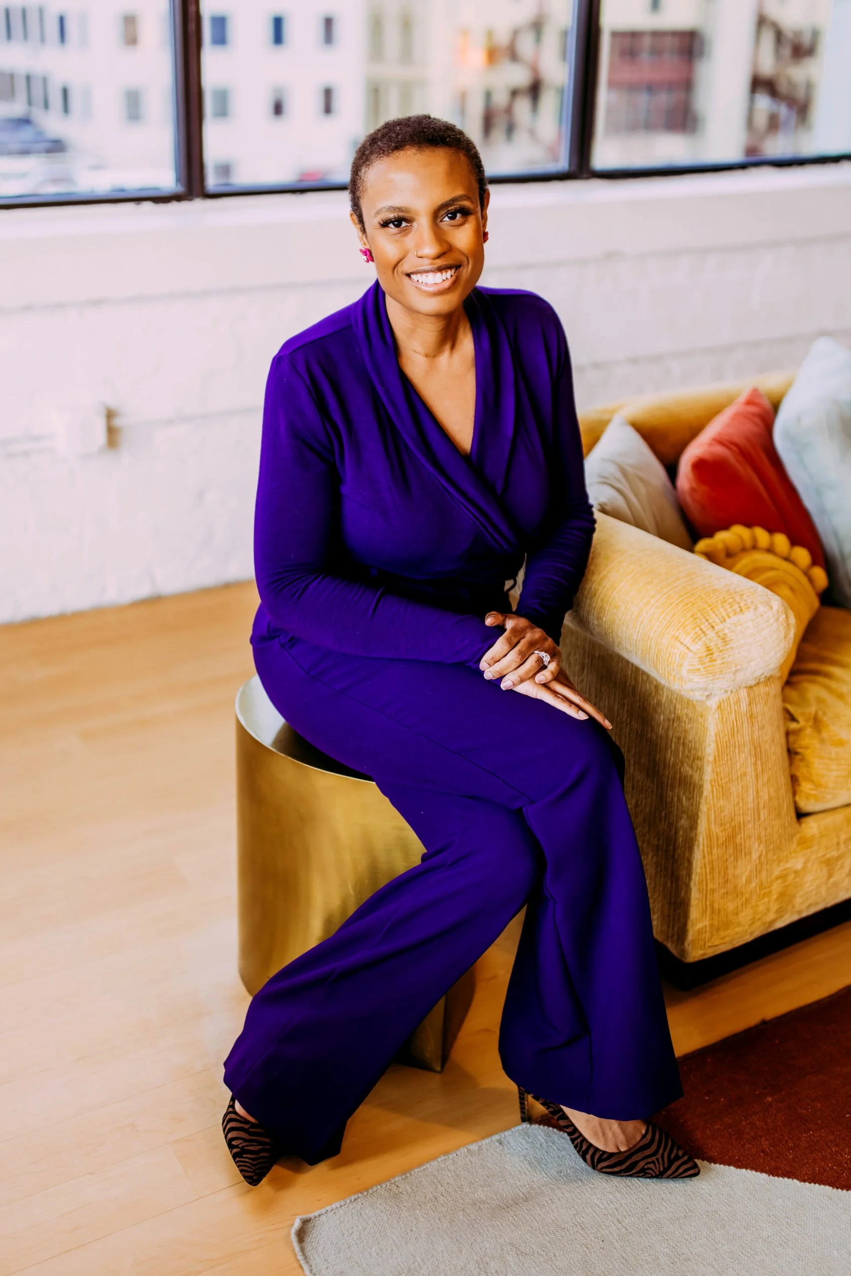 A woman with short hair, dressed in a purple outfit and zebra-print heels, sitting on a gold-colored stool in a brightly lit room with a large window, colorful pillows on a yellow couch, and a cityscape view outside.