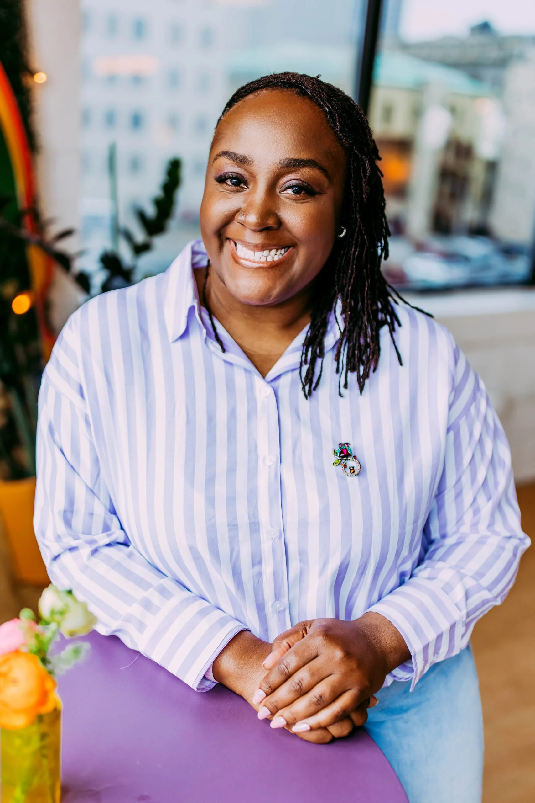 A woman with dark skin and dreadlocks smiling at the camera, wearing a light blue and white striped button-up shirt with a colorful bear pin, standing indoors near a window with a cityscape view.