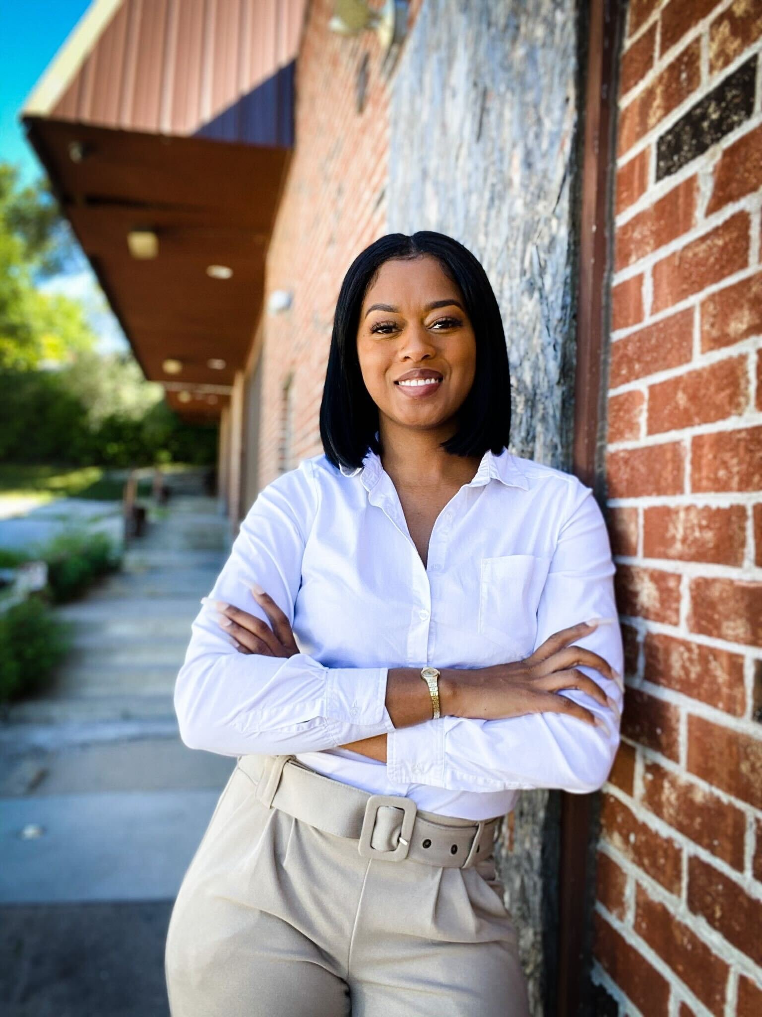 A woman with dark hair, wearing a white blouse and beige pants, standing outdoors against a brick wall, with her arms crossed and smiling.