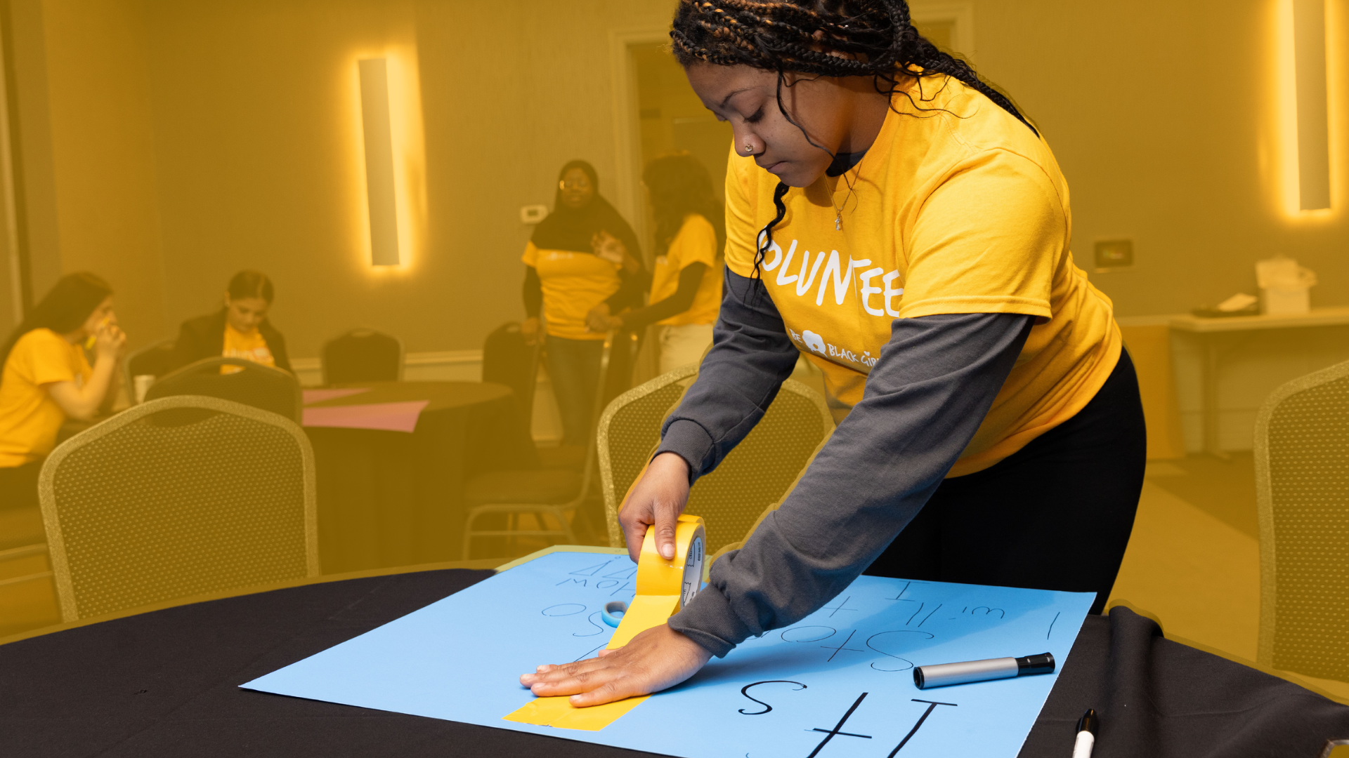 A woman in a yellow volunteer shirt is taping a blue poster board with writing on it. She is leaning over the table, focused on her task. In the background, other volunteers in yellow shirts are sitting and standing in a room with yellow walls.