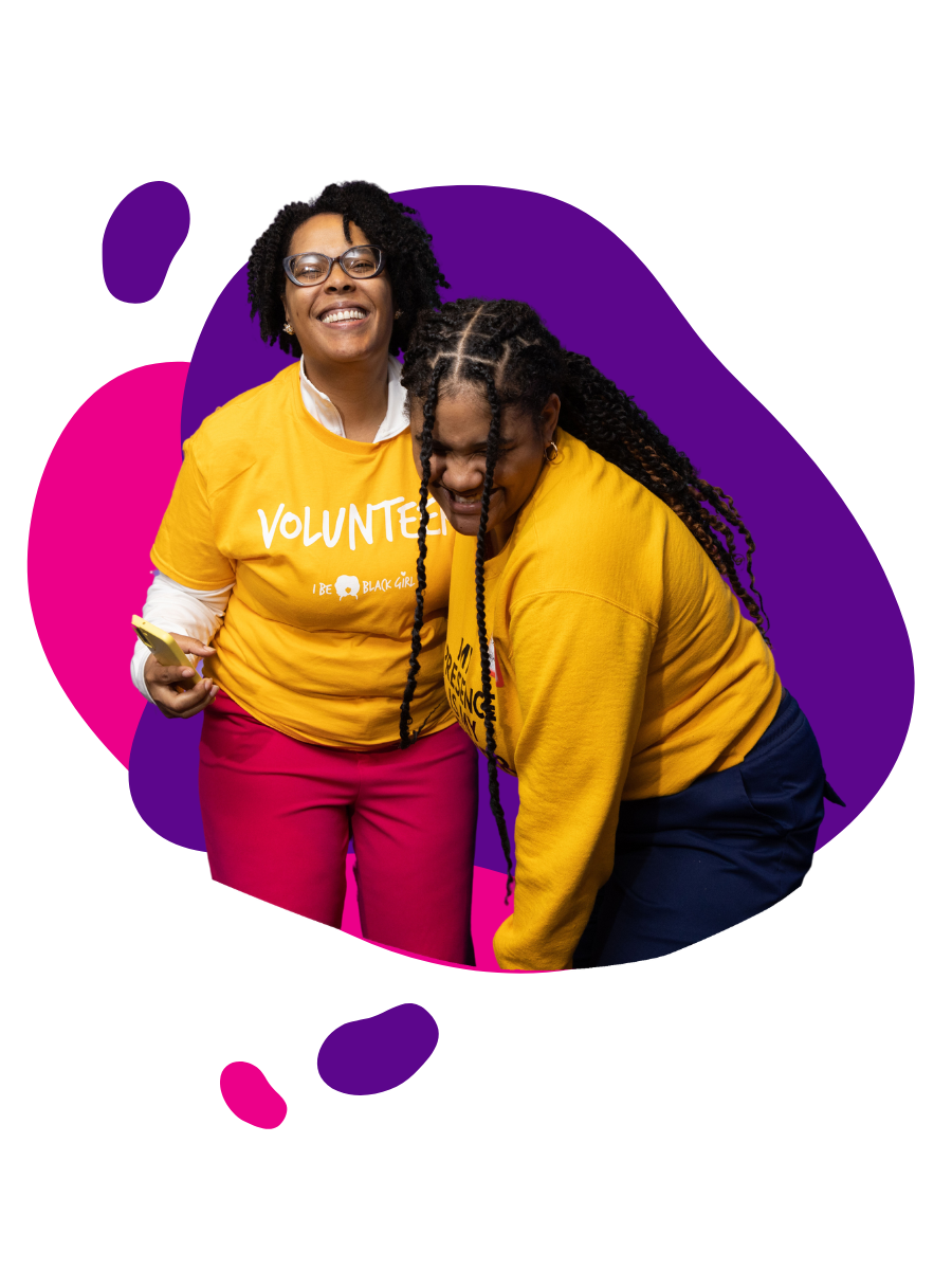 Two women wearing yellow volunteer shirts are smiling and laughing together. One woman has glasses and short curly hair, holding a phone in her left hand. The other woman has long braided hair. They are against a colorful background with pink and purple elements.