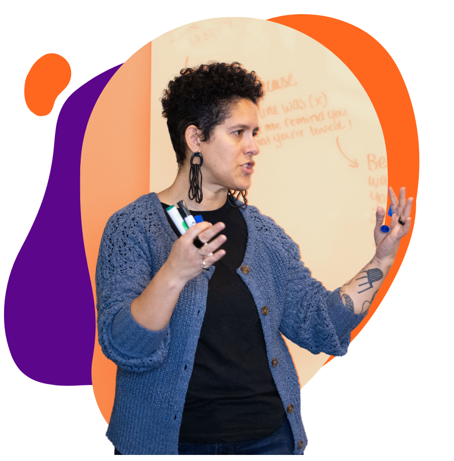 A woman with short curly hair and earrings speaking and gesturing with her hands in front of a whiteboard with orange writing.
