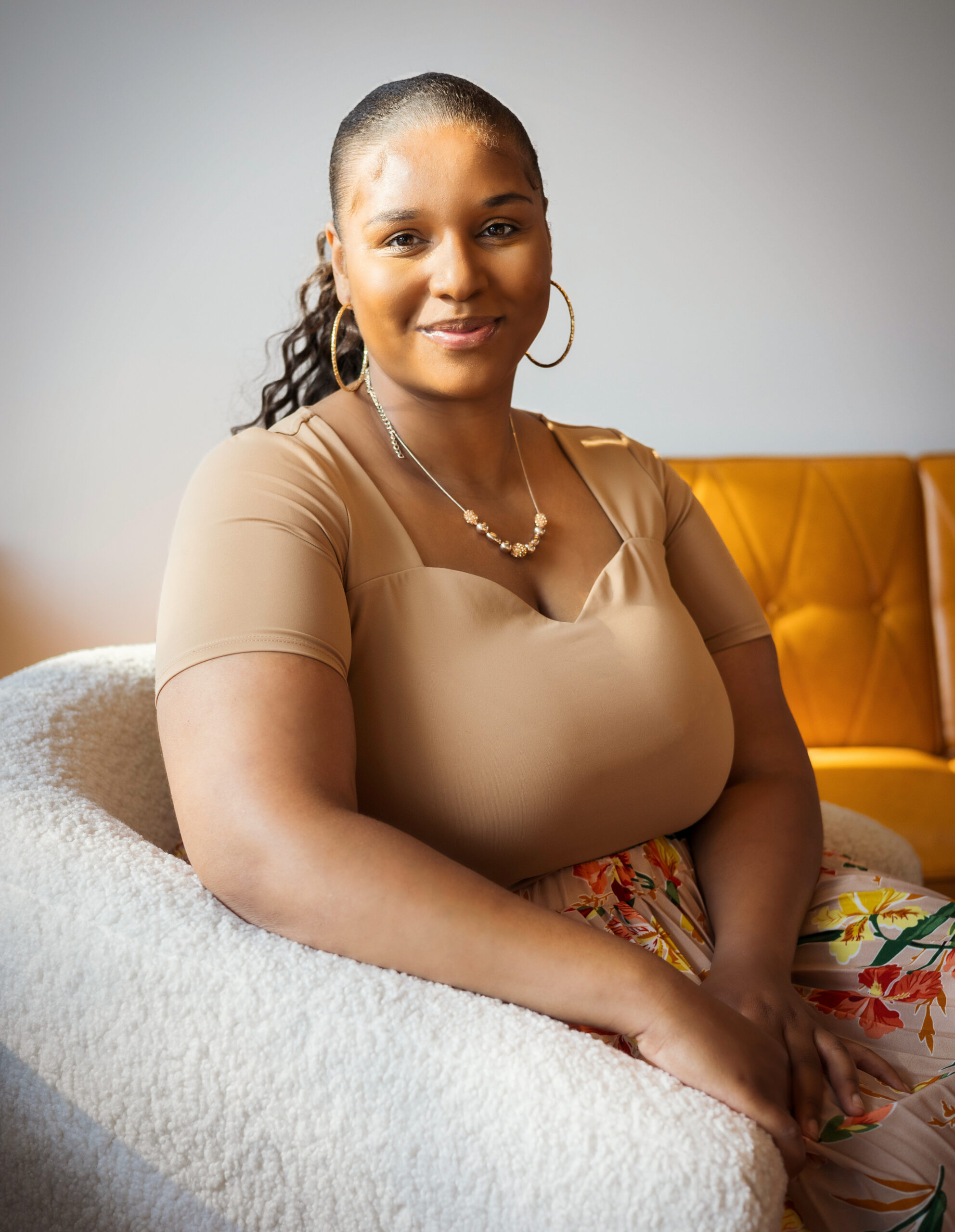 A smiling woman with dark hair pulled back, wearing gold hoop earrings, a gold beaded necklace, a beige top, and floral pants, sitting on a plush white chair with a brown leather couch in the background.