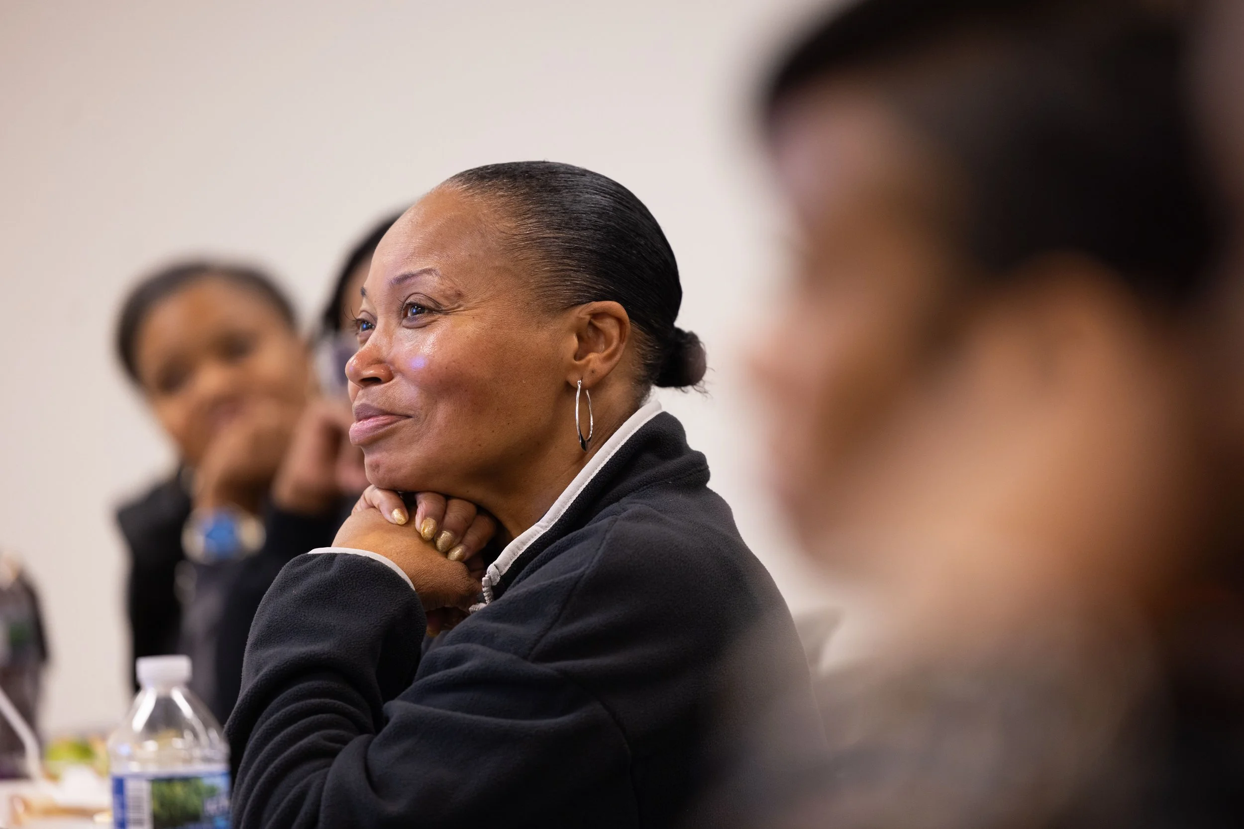 A woman with her hair in a bun, wearing hoop earrings, sitting with her hands under her chin, smiling and listening attentively during a discussion or meeting surrounded by other women.