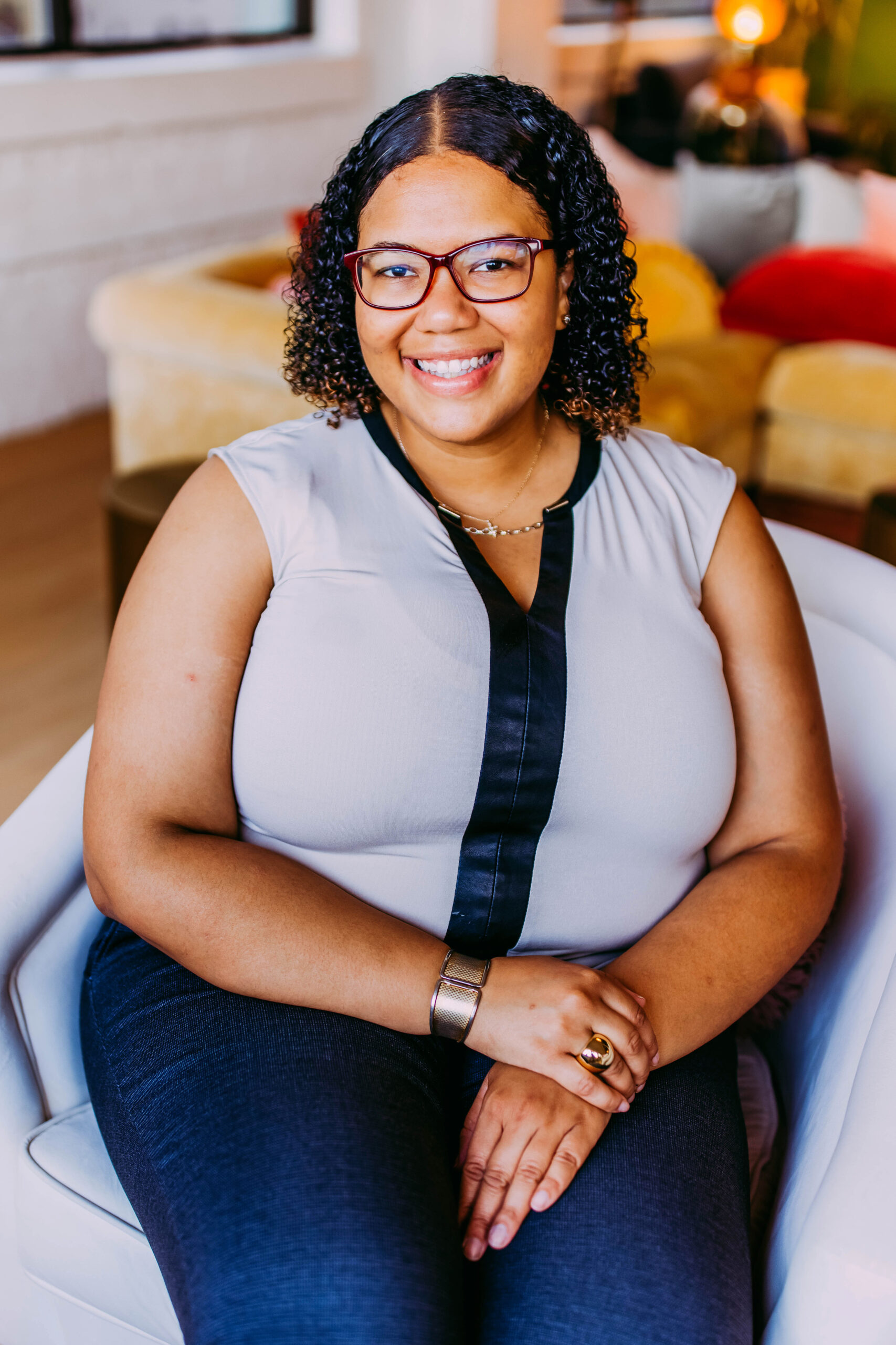 A smiling woman with glasses, curly hair, wearing a sleeveless gray top with black accents, sitting on a white chair in a colorful indoor setting.
