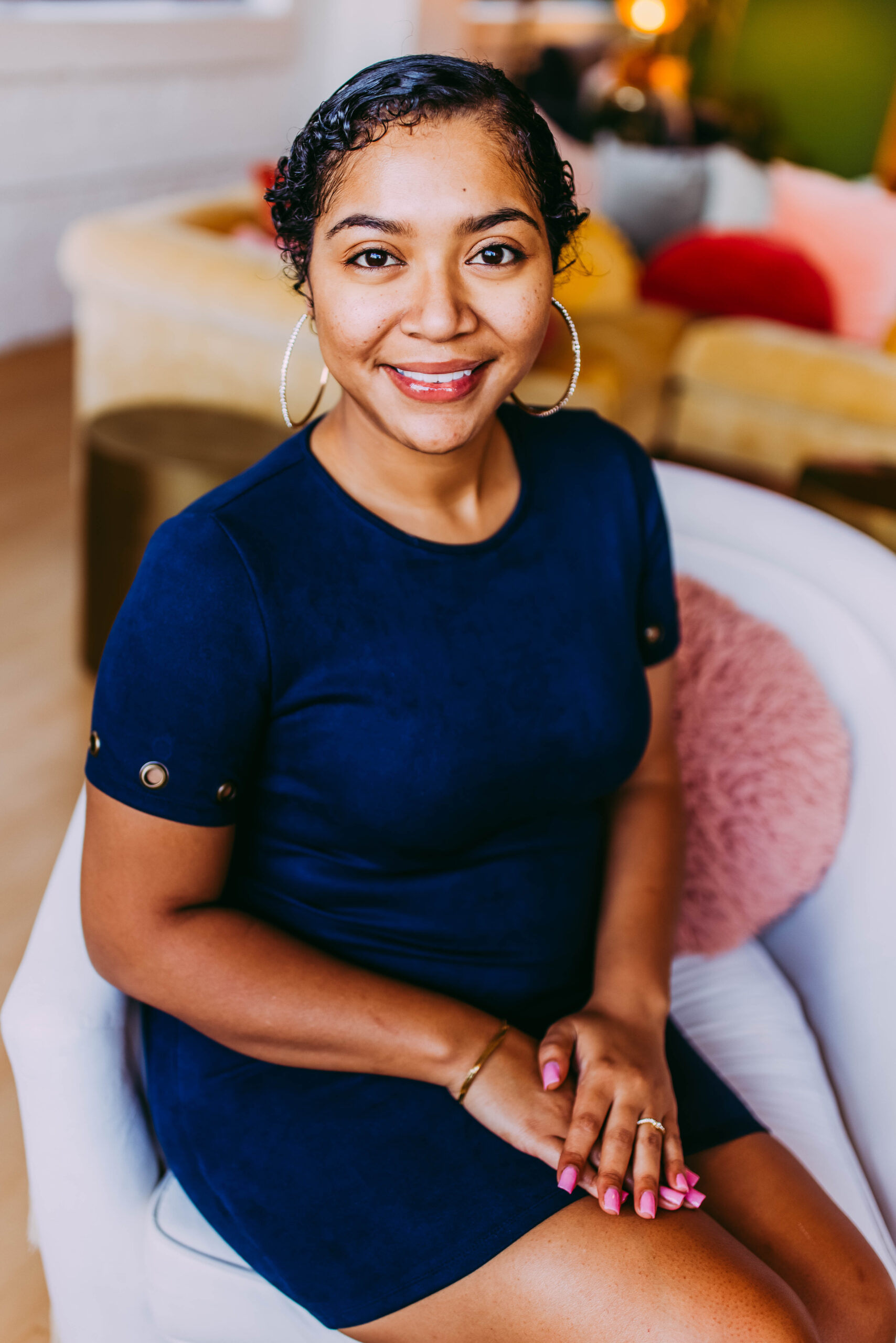A woman sitting in a white armchair, smiling at the camera in a cozy living room with colorful cushions and a yellow sofa in the background.