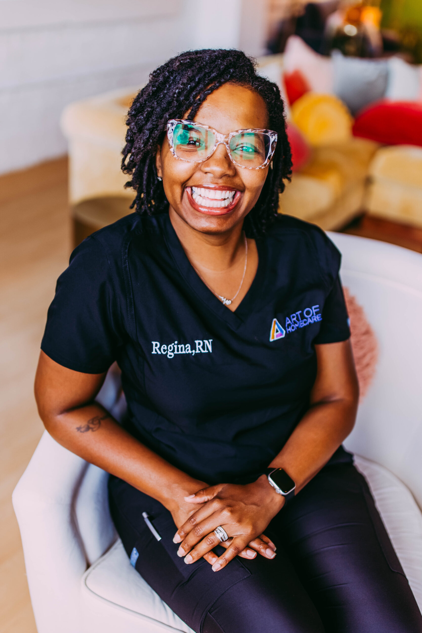 A smiling woman wearing glasses and a black medical uniform with the name tag 'Regina, RN', sitting on a white chair in a cozy, colorful living room.