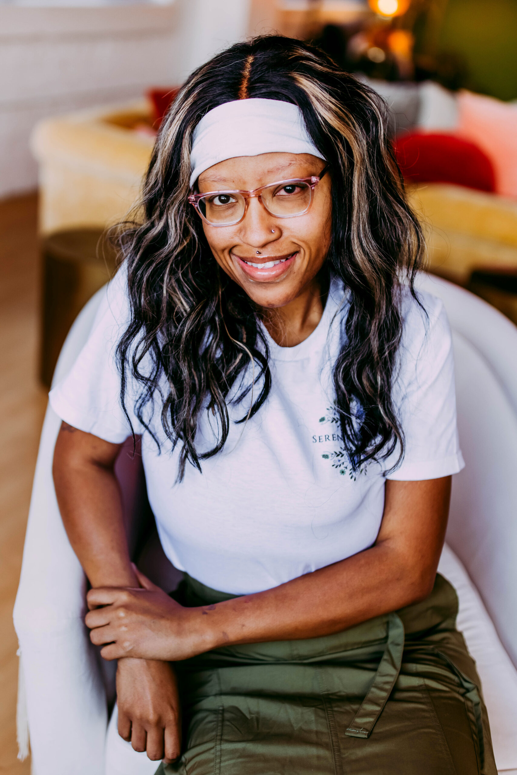 A woman with curly dark hair streaked with blonde highlights, wearing glasses, a white headband, and a white t-shirt, sitting on a white chair in a cozy indoor setting with a soft smile.