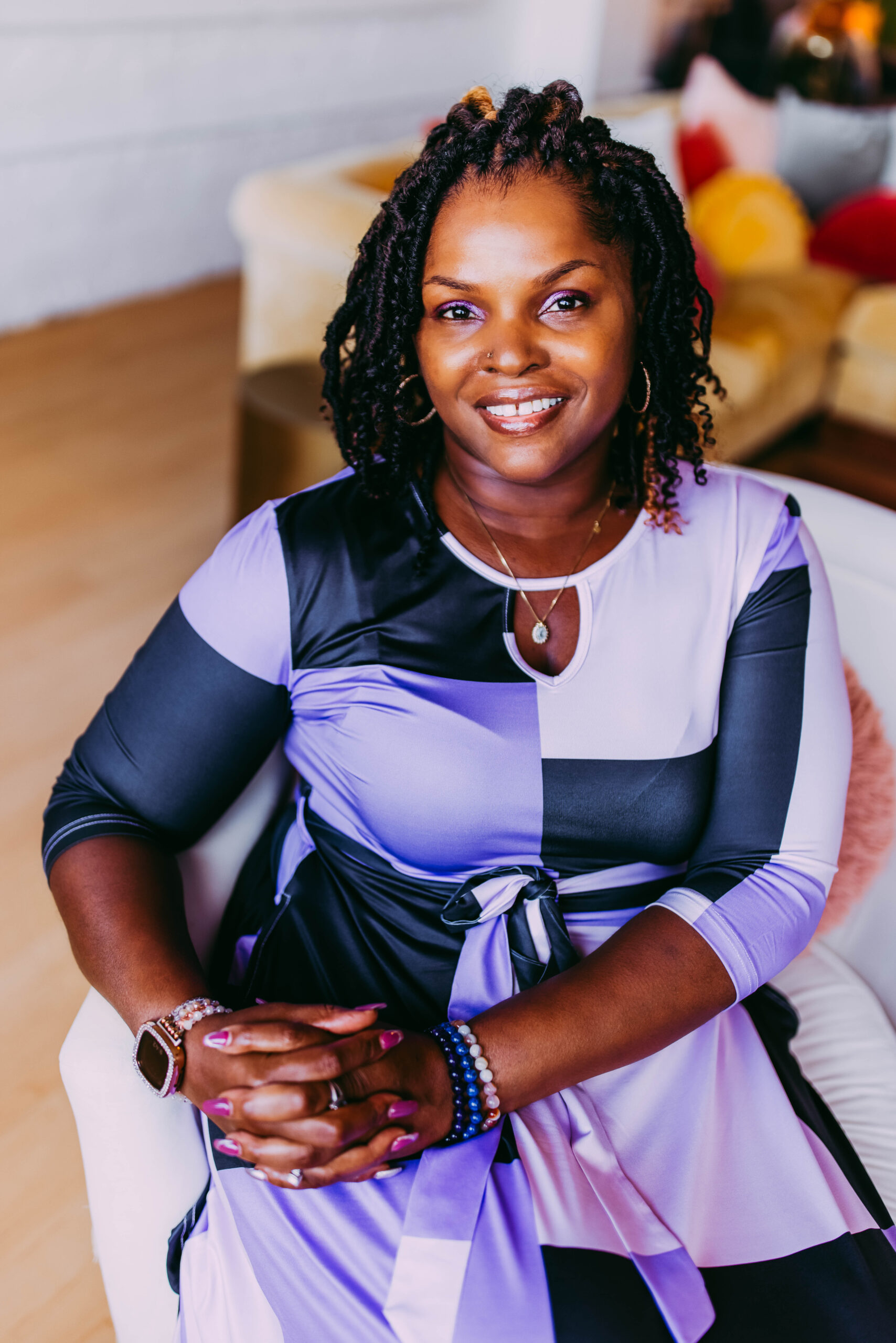 A woman with braided hair and a nose piercing sitting indoors, smiling, wearing a color-blocked purple, black, and white dress, with jewelry and bracelets.
