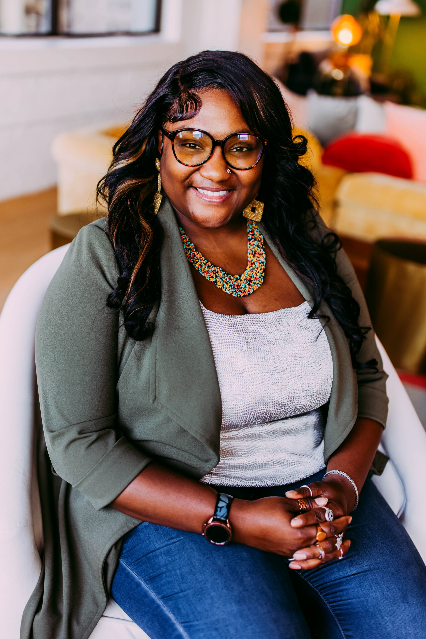 A smiling woman with long dark hair, glasses, and colorful jewelry sitting in a cozy room with colorful cushions and warm lighting.