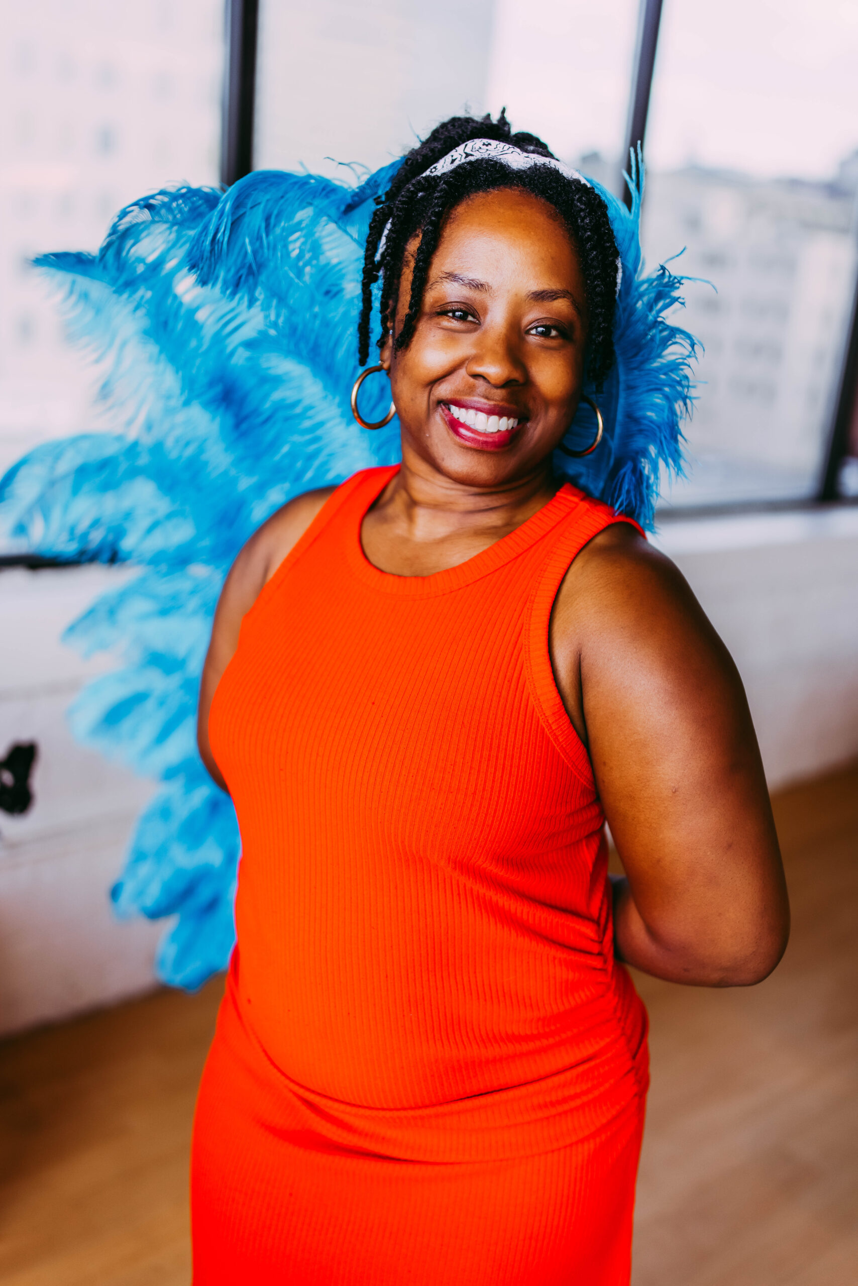 A woman smiling, wearing an orange sleeveless dress, with blue angel wings behind her, and a white cloth headband, standing indoors with windows in the background.