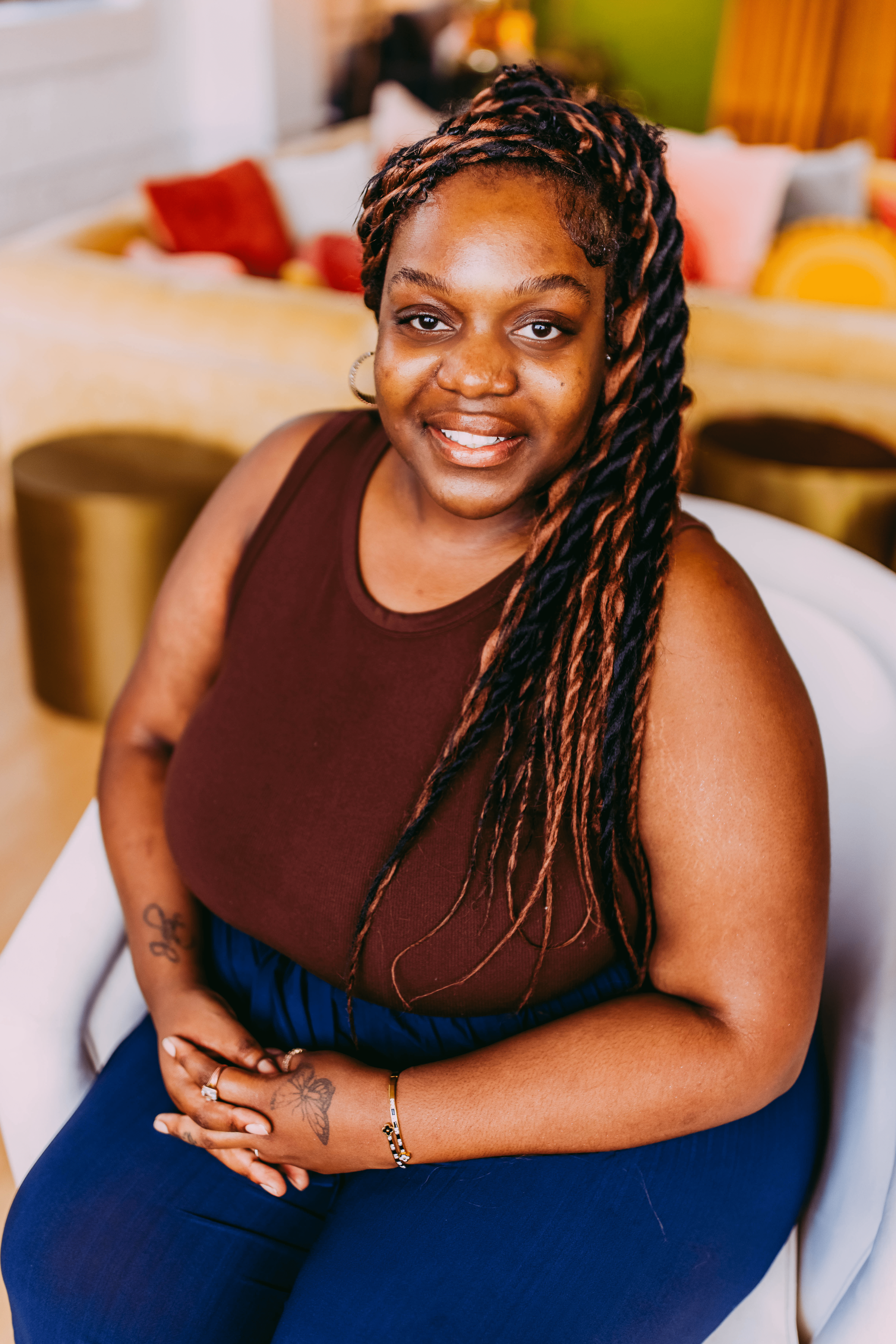 A woman with long, twisted braids sitting indoors, wearing a sleeveless maroon top and dark blue pants, smiling with her hands clasped on her lap.