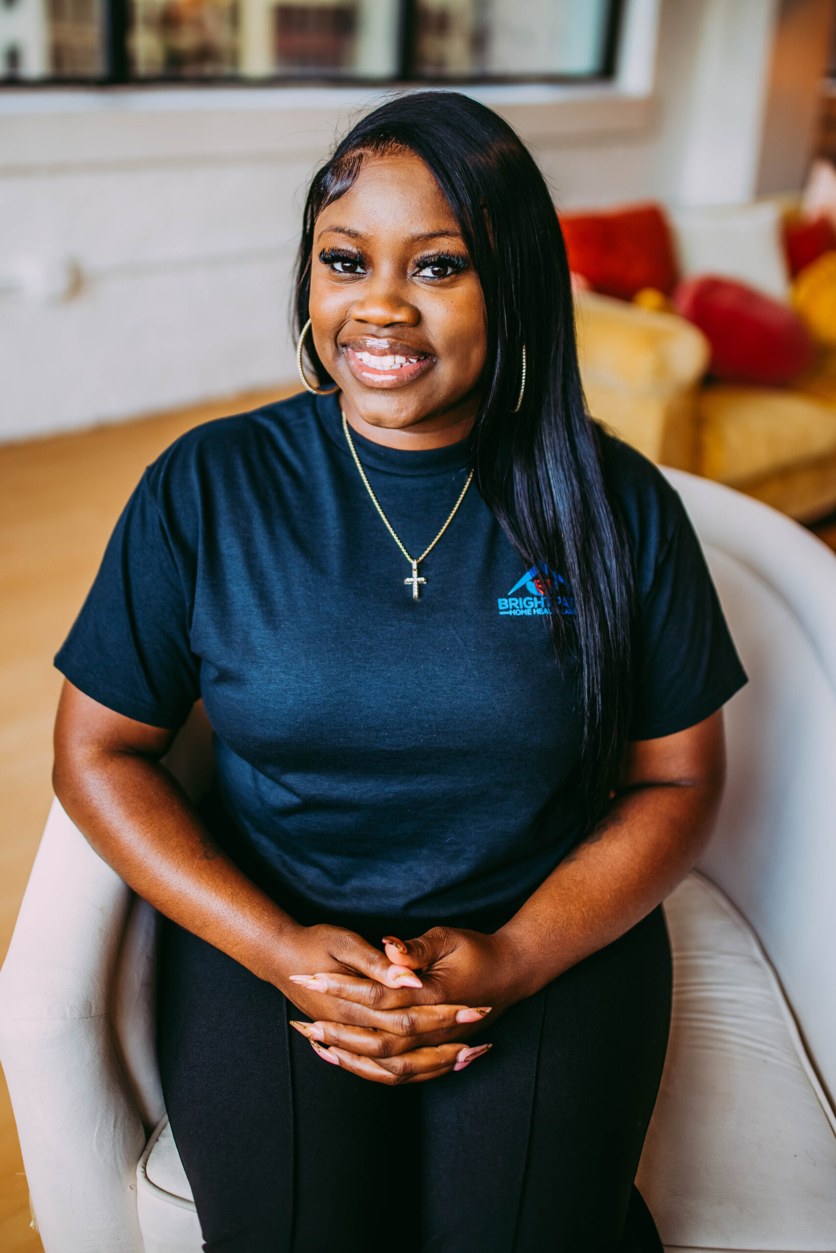 A woman with long black hair, wearing hoop earrings, a cross necklace, and a navy blue T-shirt, sitting on a white armchair and smiling at the camera.