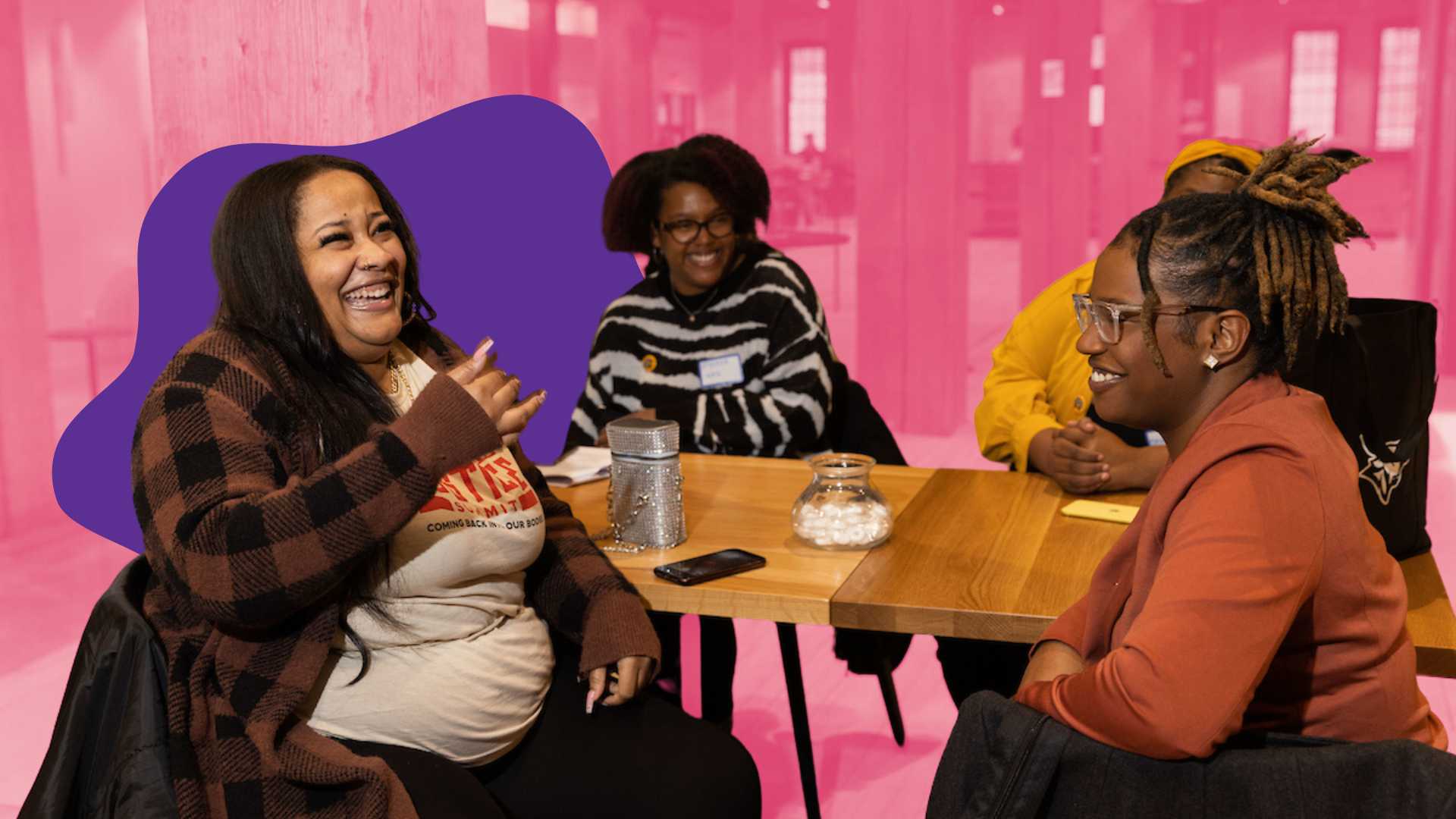 Four women sitting at a wooden table, laughing and engaging in conversation. The background is pink with a semi-transparent overlay, and the table has a glass jar with white objects, a phone, and containers.