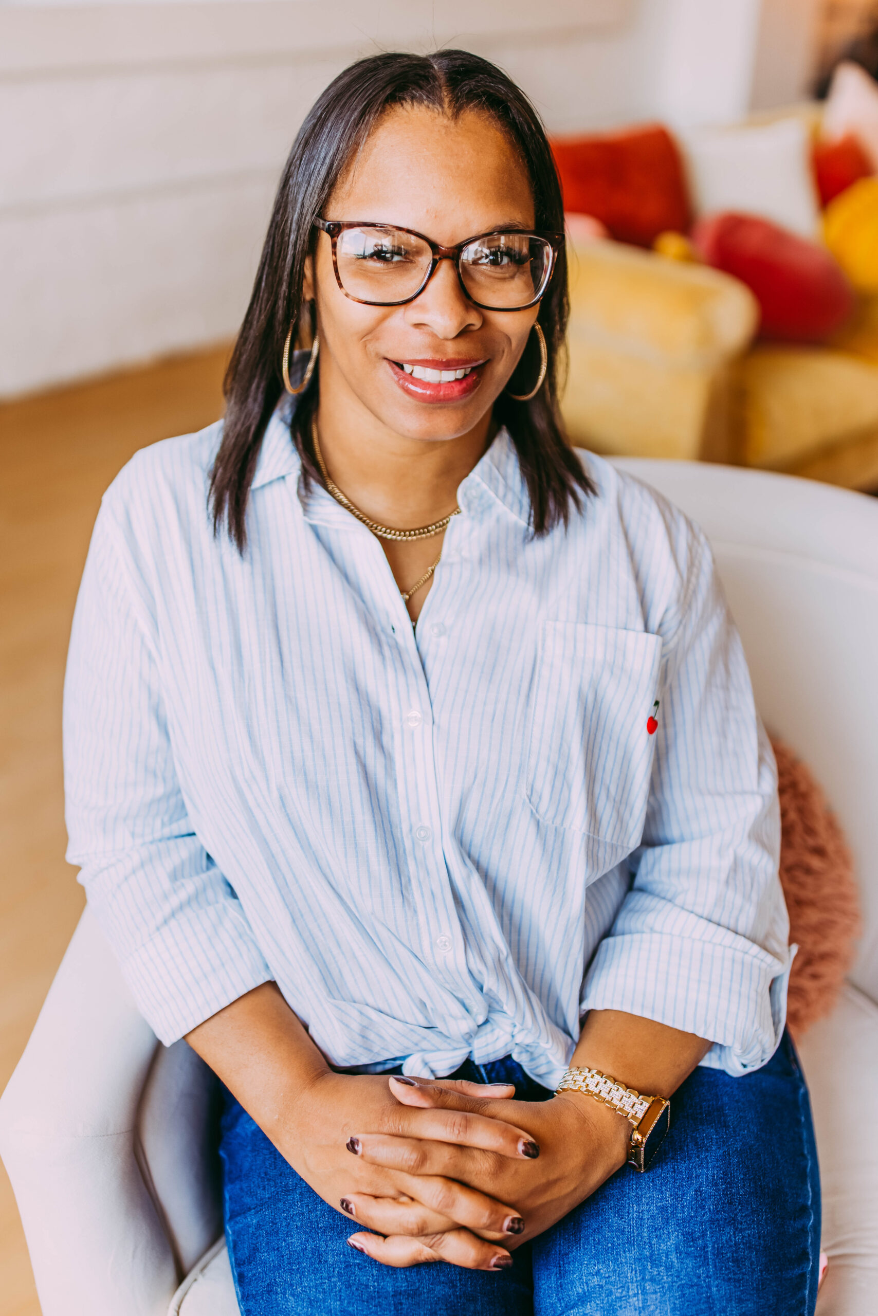 A smiling woman wearing glasses, a white striped shirt tied at the waist, blue jeans, and gold jewelry, sitting on a white chair in a brightly lit room with colorful pillows in the background.