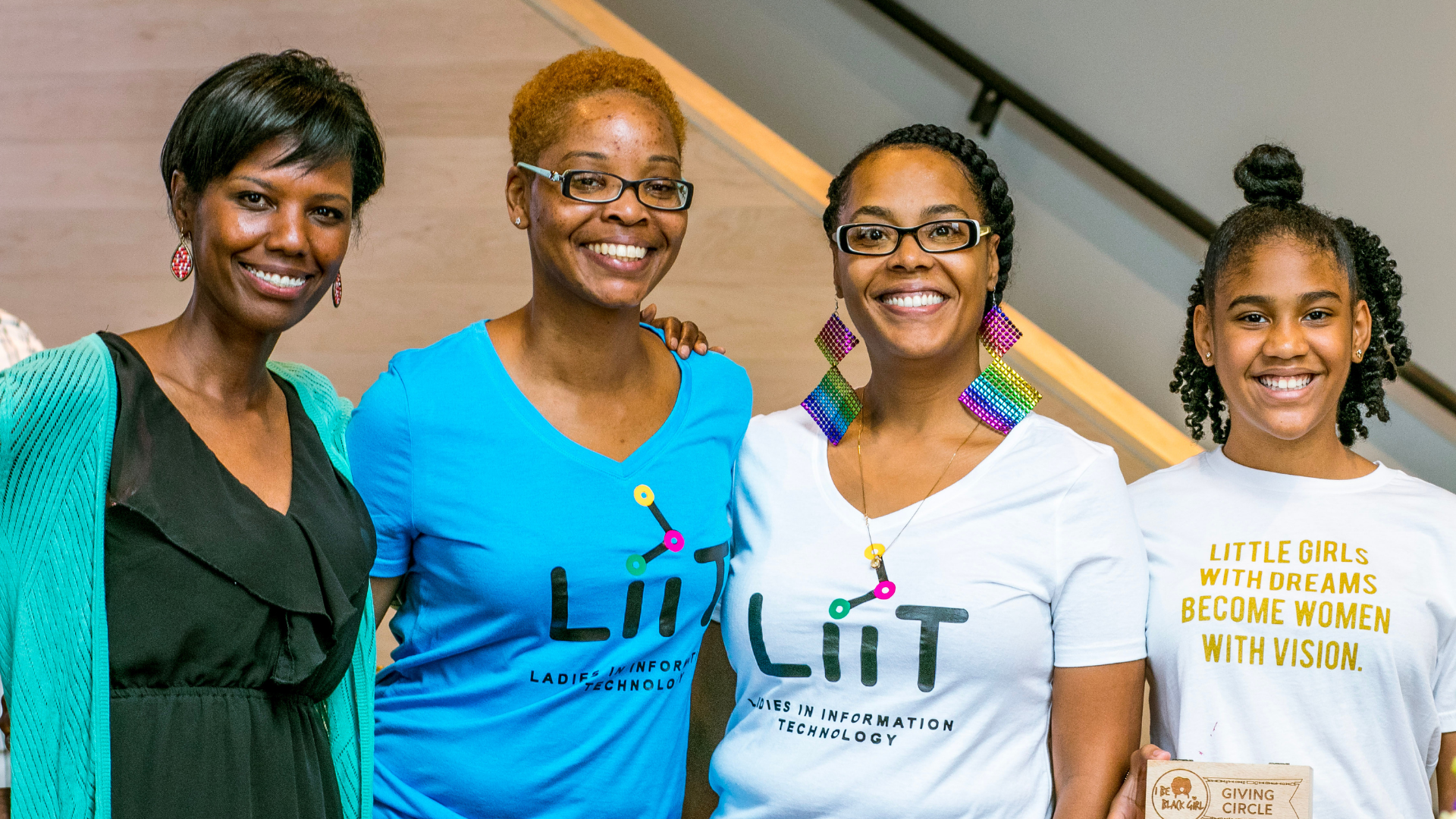 Four women standing together, smiling at the camera. Two women are wearing white t-shirts with the 'LiiT' logo and text about women in technology, one woman is wearing a blue t-shirt with the same logo, and the fourth woman is wearing a black dress with a turquoise cardigan. The woman on the far right has a name tag and a shirt with a quote about girls with vision.