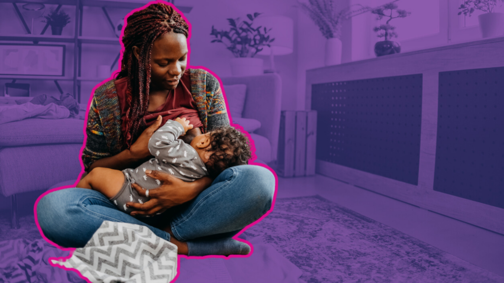 A woman with braided hair sitting cross-legged on the floor, breastfeeding a baby in a cozy living room